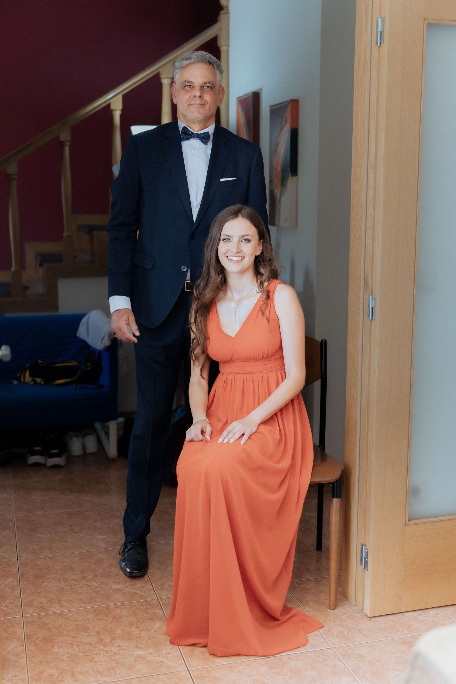 noivo de smoking preto está ao lado da noiva sentada em um corredor interno, vestindo um elegante vestido coral. Casamento em Esposende.