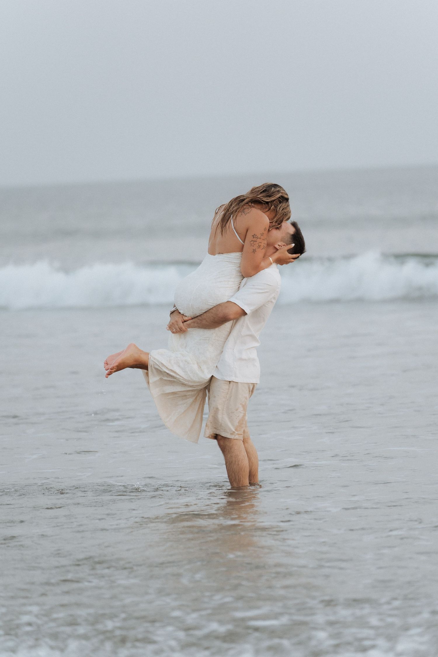 Casal romântico se abraça de forma divertida enquanto está com os pés na agua rasas do oceano, vestindo roupas brancas e bege.