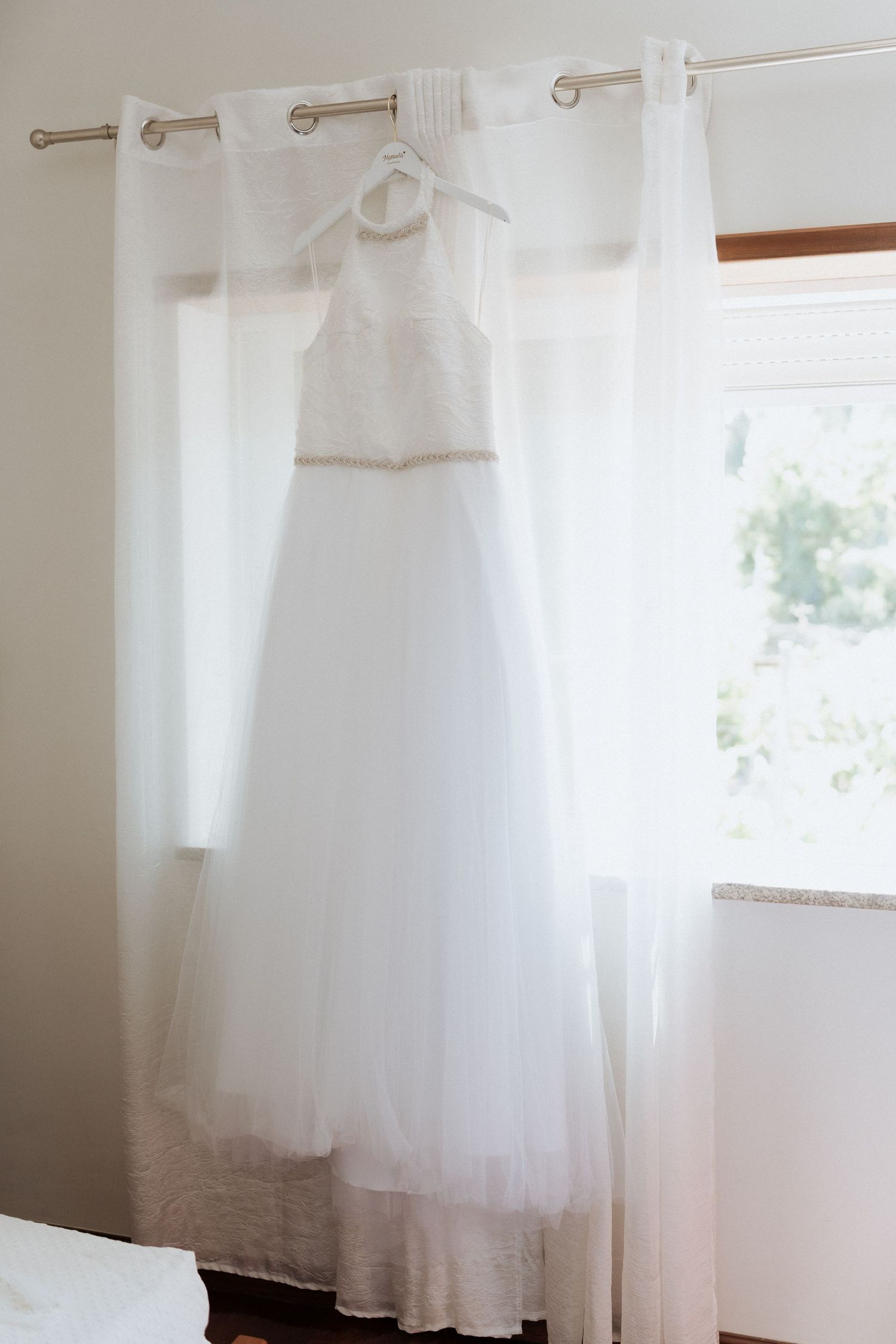 Elegante vestido de noiva branco pendurado num varão de cortina em frente a uma janela iluminada com cortinas. Fotografia de Paulo Roque.
