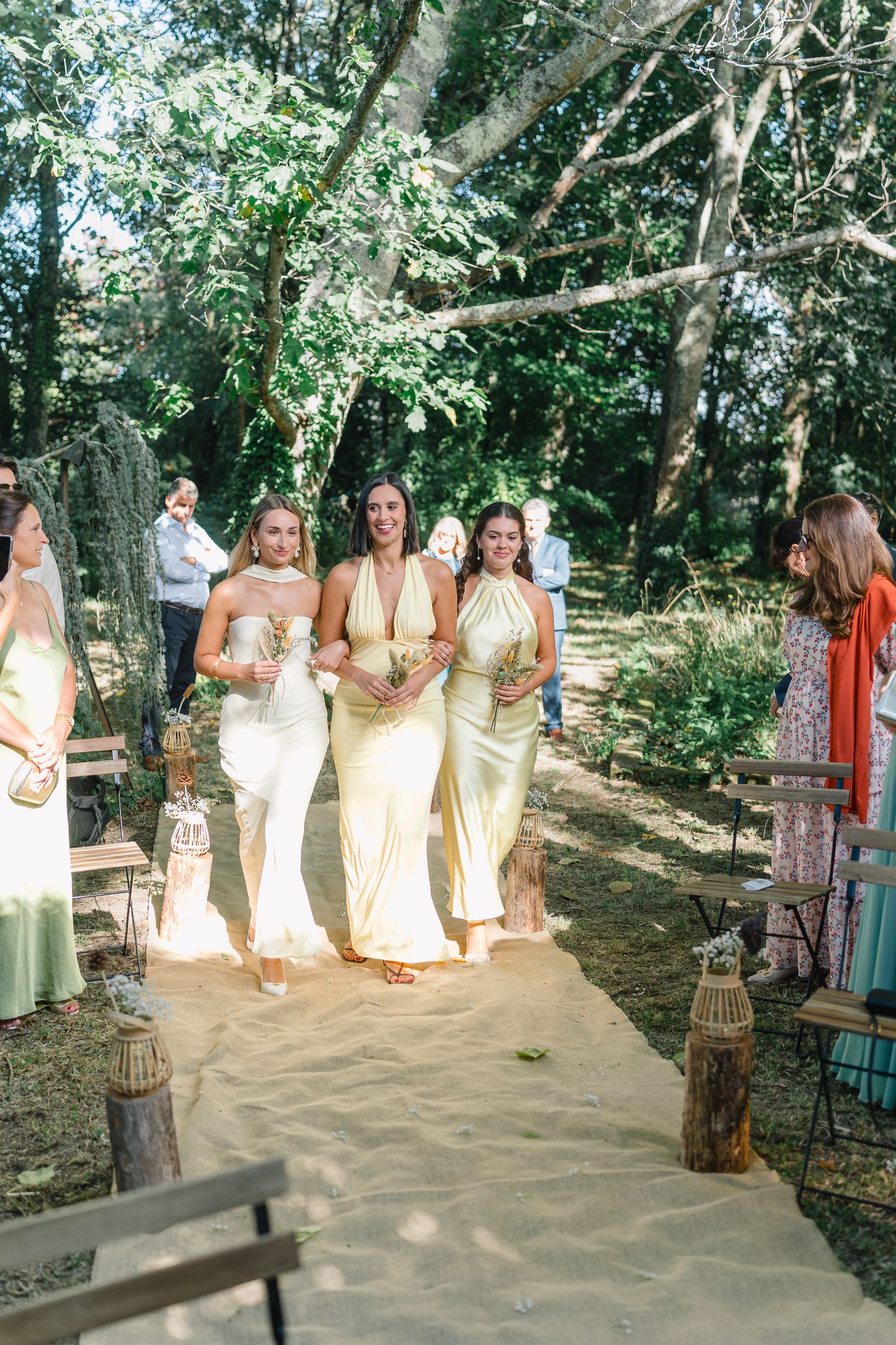 Bridesmaids in pale yellow dresses gather outdoors under trees for a wedding photo session.