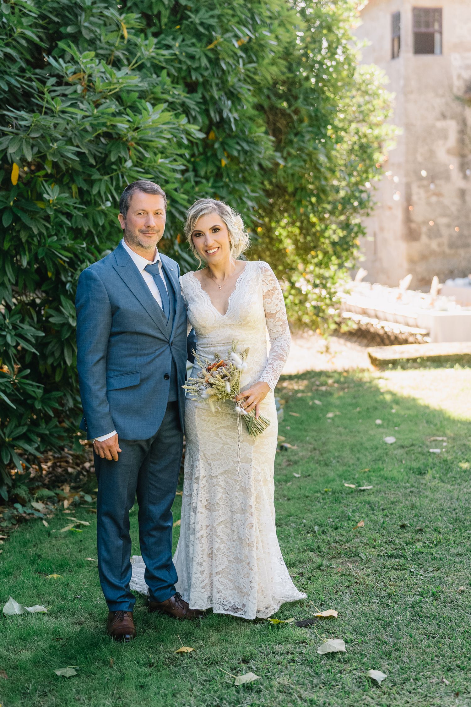 Noivos pousam juntos ao ar livre, em trajes de casamento, com uma vegetação luxuriante e um muro de pedra ao fundo. Ponte de Lima.