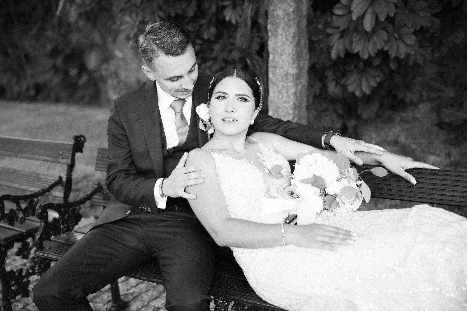 A tender black and white wedding photo of a couple relaxing on a garden bench with flowers.