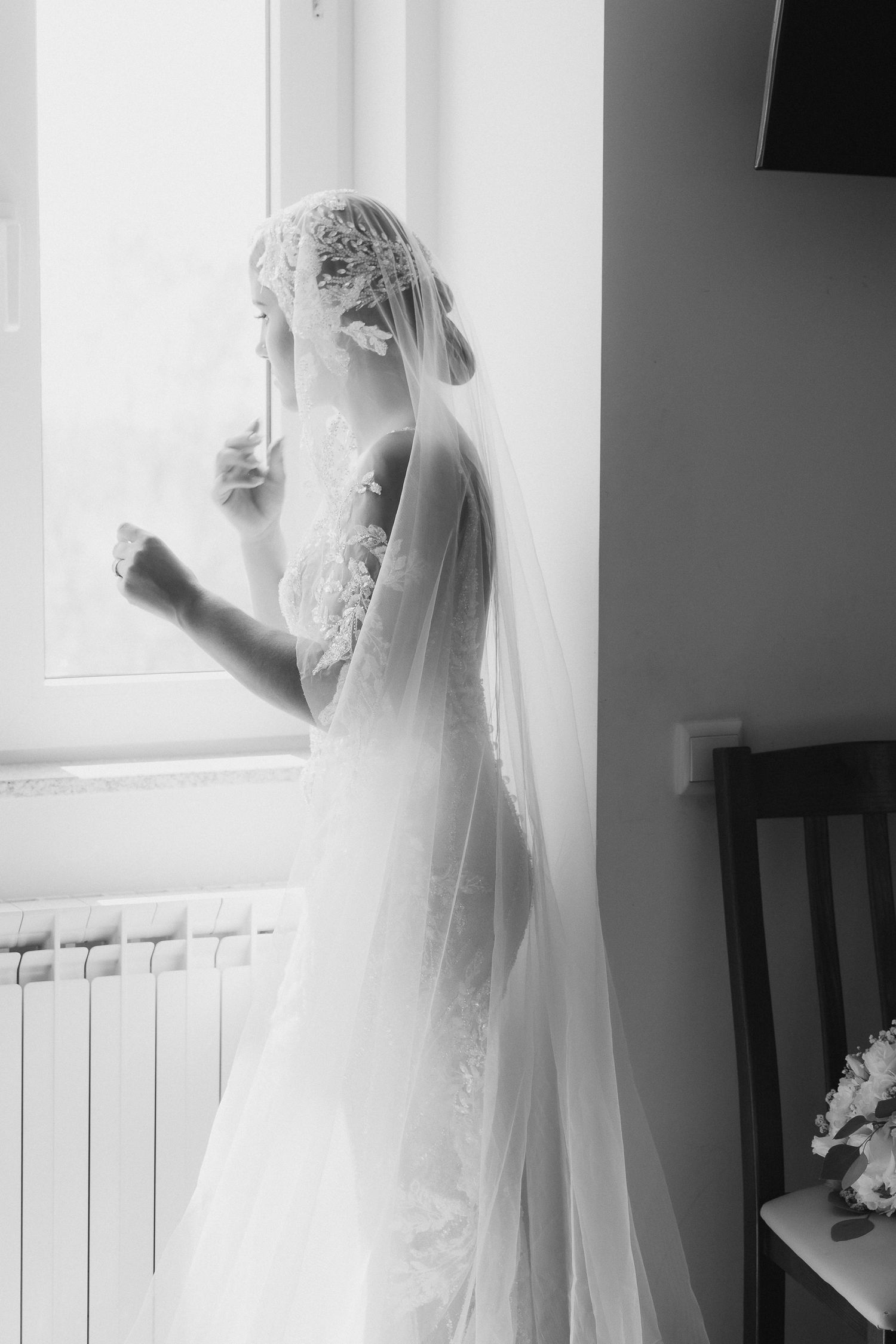 A bride in a flowing white wedding dress and veil stands silhouetted by bright window light in a dreamy black and white photo.