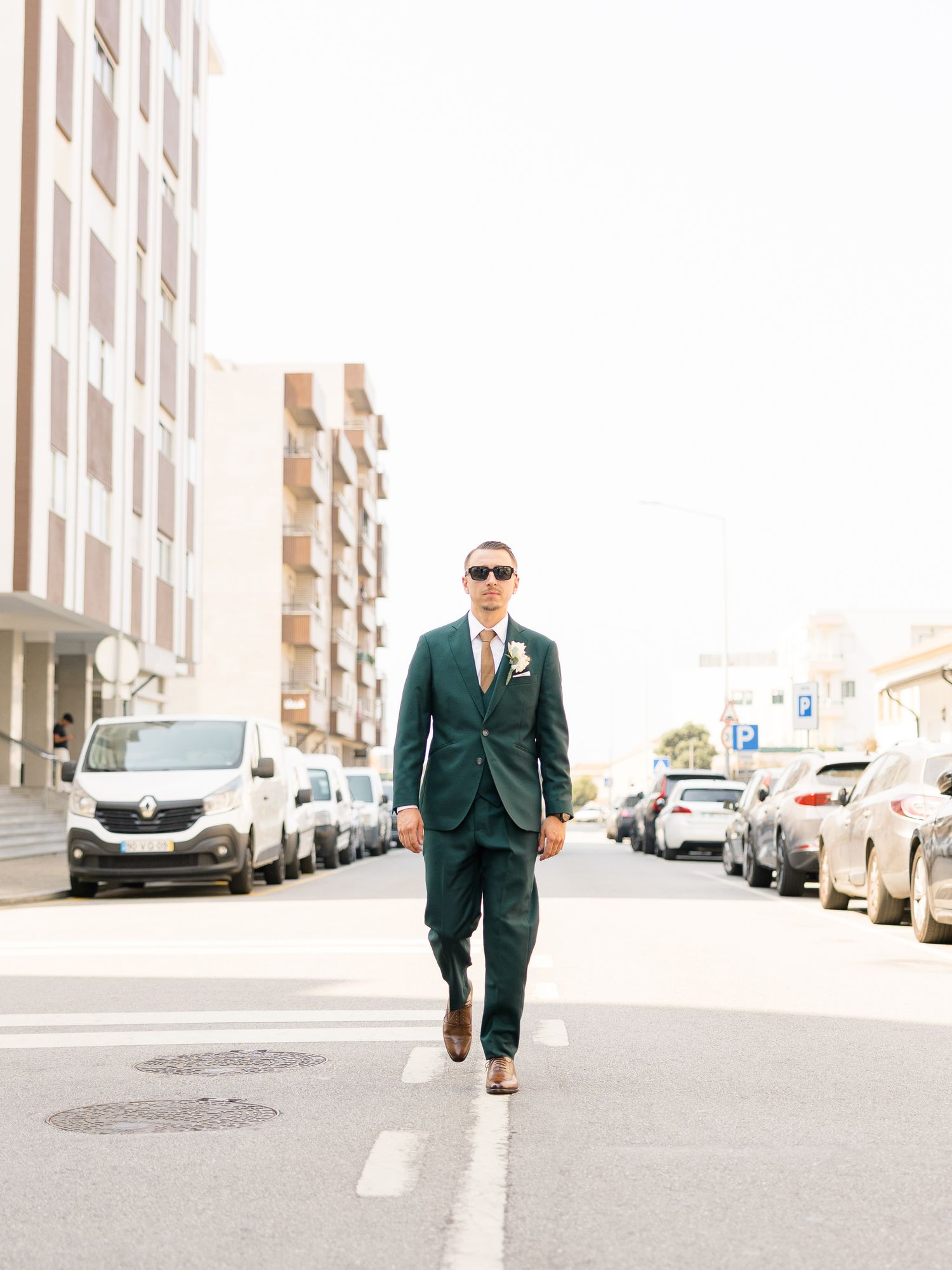 Man in dark suit and sunglasses walks confidently down an urban street lined with parked cars.
