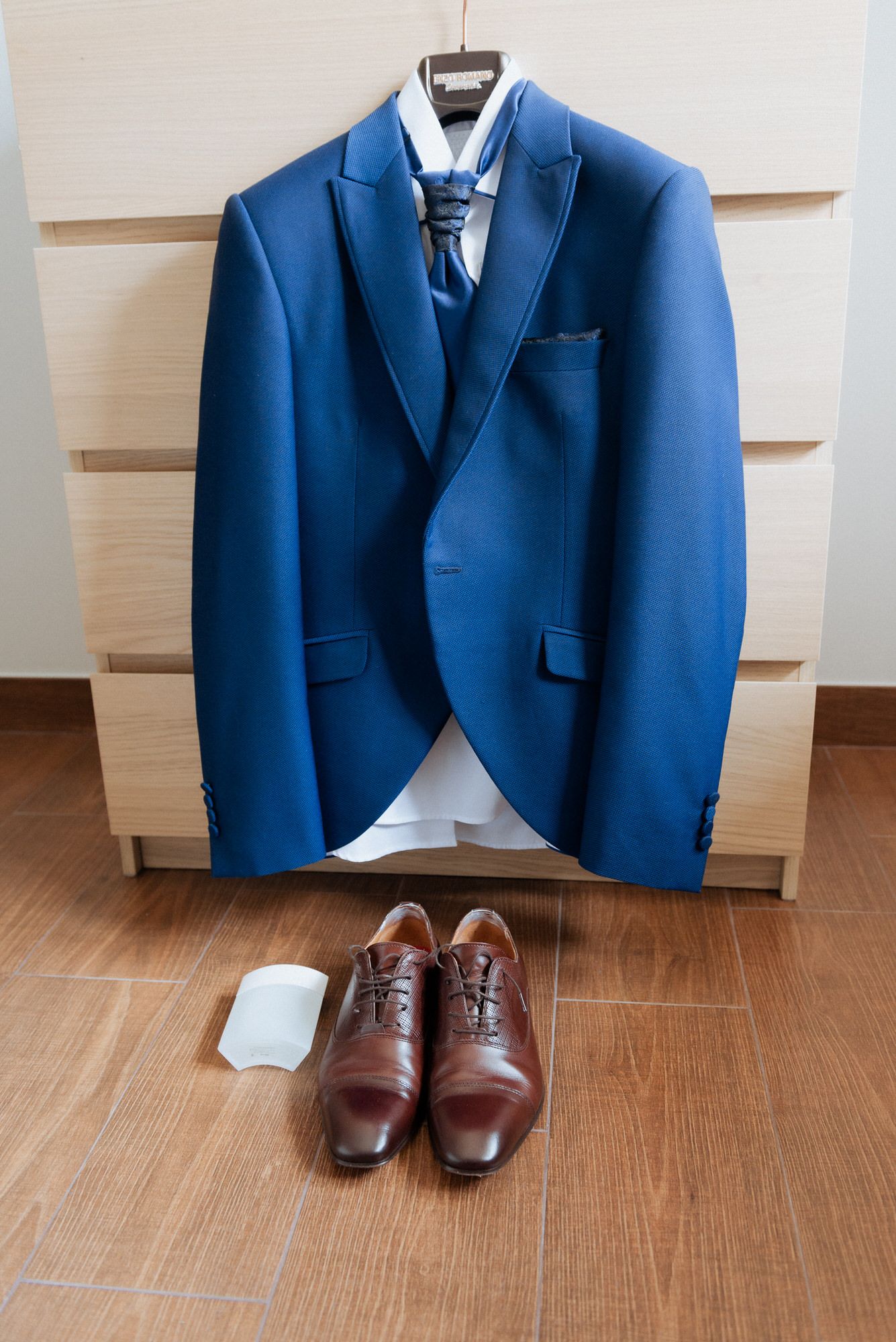 A bright blue suit jacket hangs with a tie and white shirt above brown leather dress shoes on hardwood flooring.