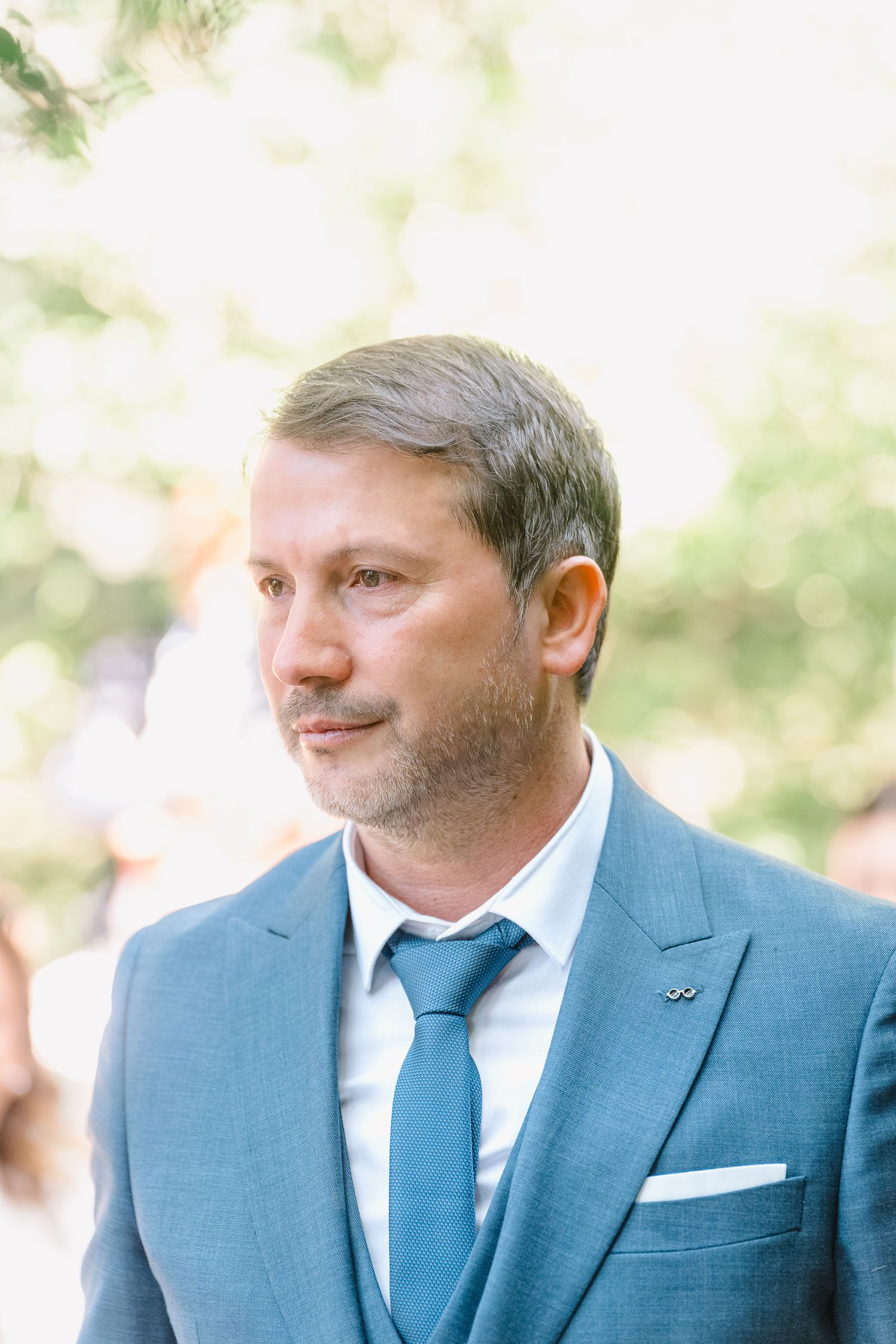 Man wearing a light blue suit and blue tie smiles while looking to the side in an outdoor setting.
