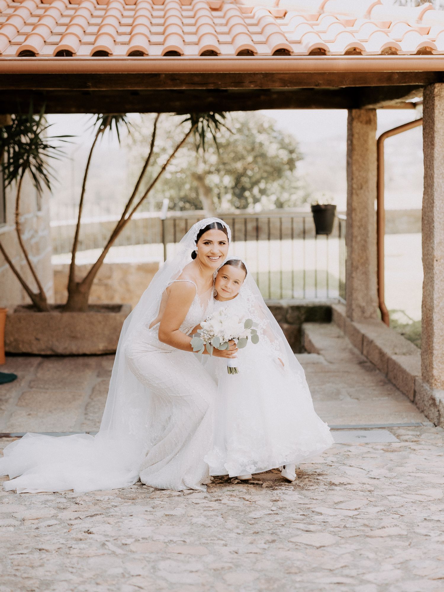 Two figures in elegant white wedding dresses share a tender moment under a covered porch with terracotta roof tiles.