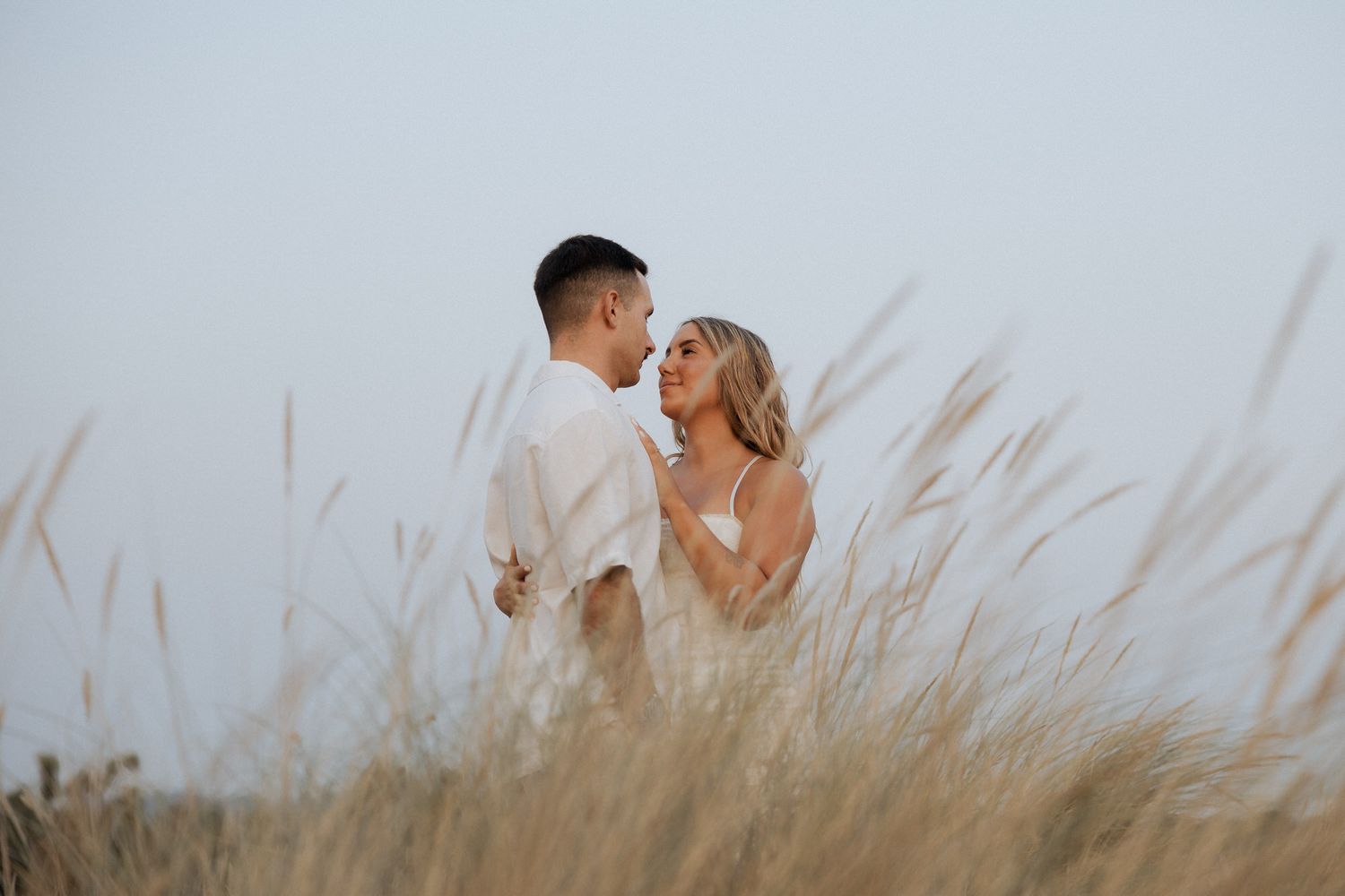 A couple shares a romantic moment in tall grass at sunset while wearing casual attire.