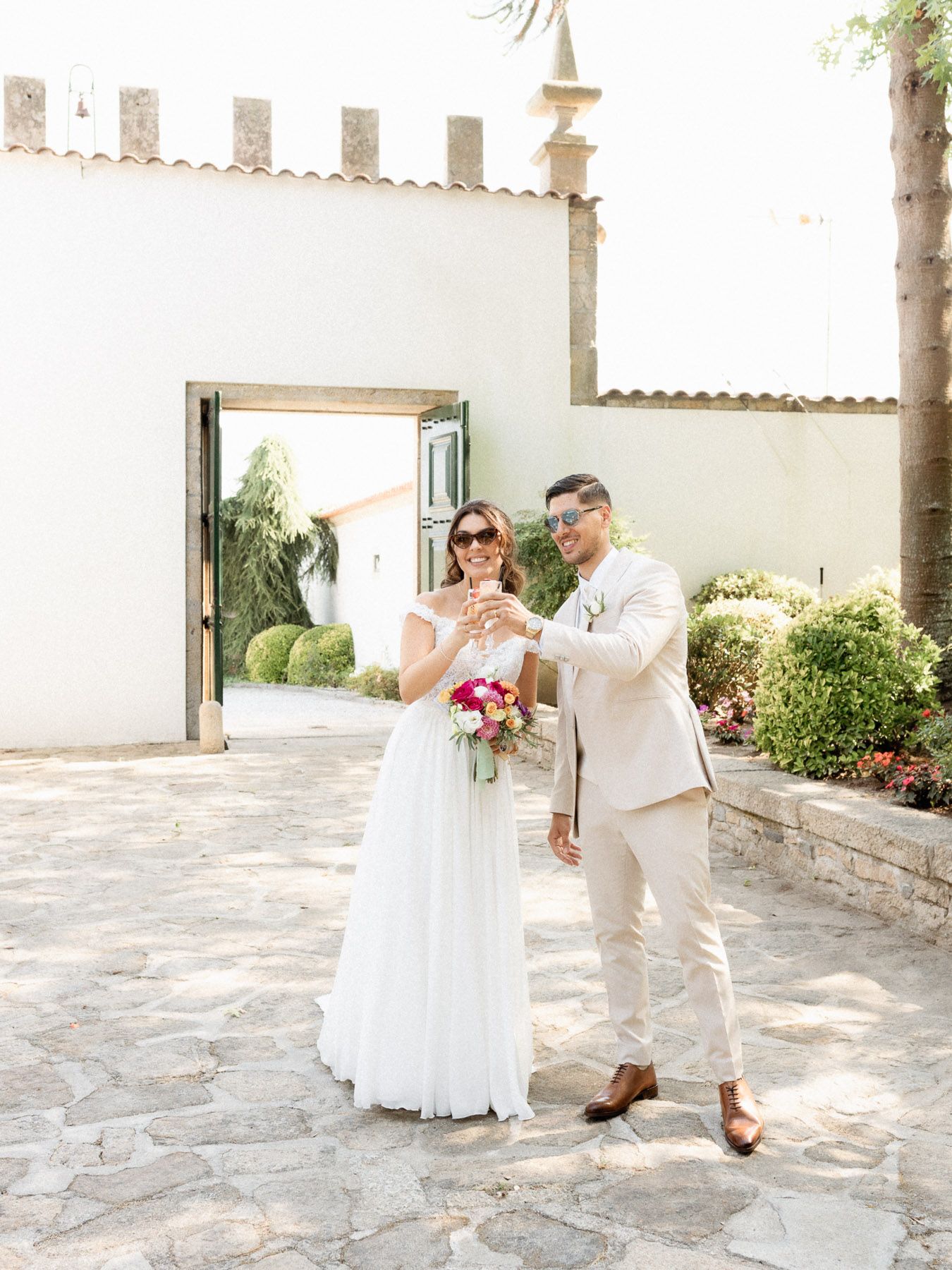 Bride and groom toasting with champagne outside a white stone venue, bride holding a pink bouquet in a white gown.
