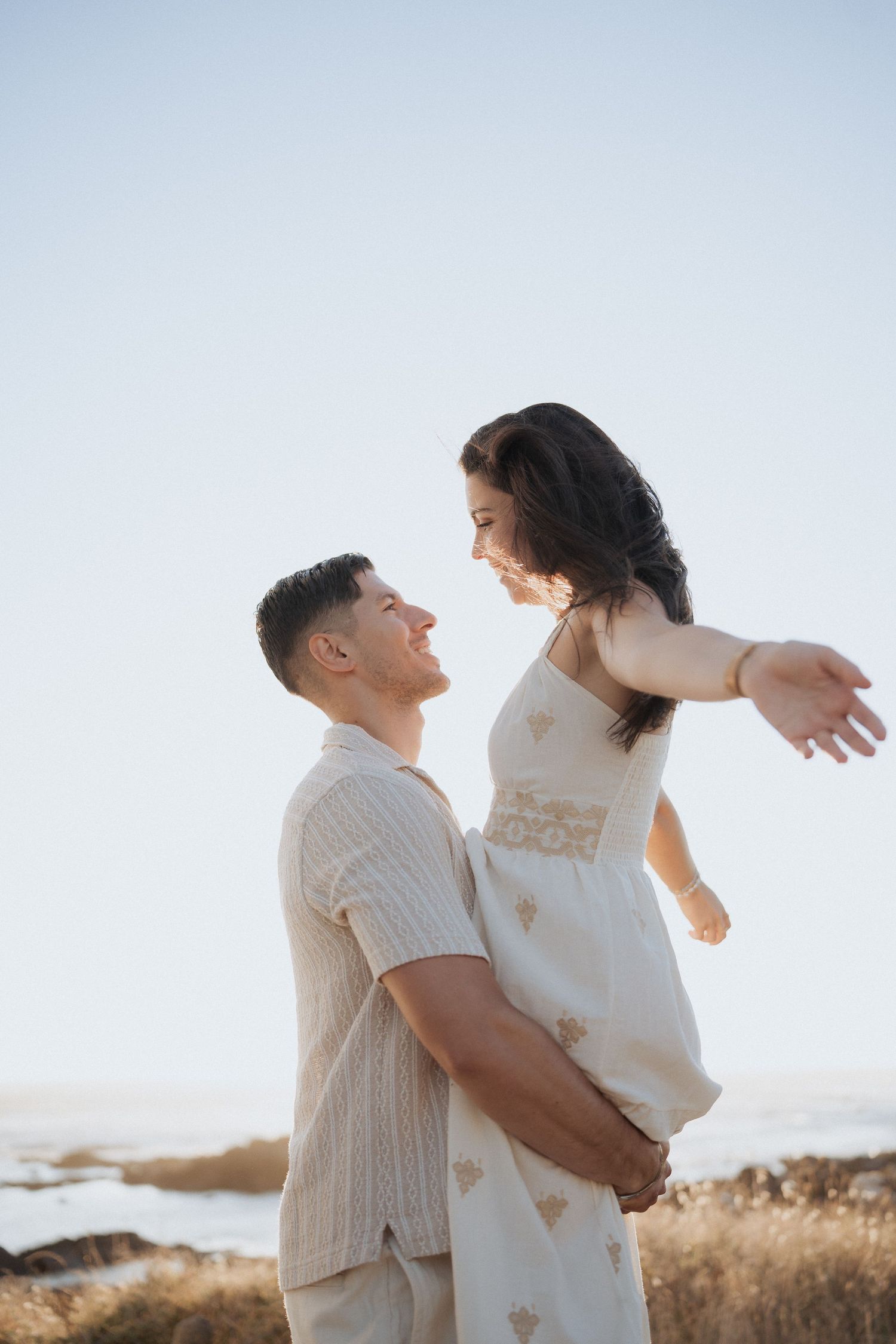 Um casal vestido de branco compartilha um momento divertido sob um céu claro. sessão fotográfica na praia