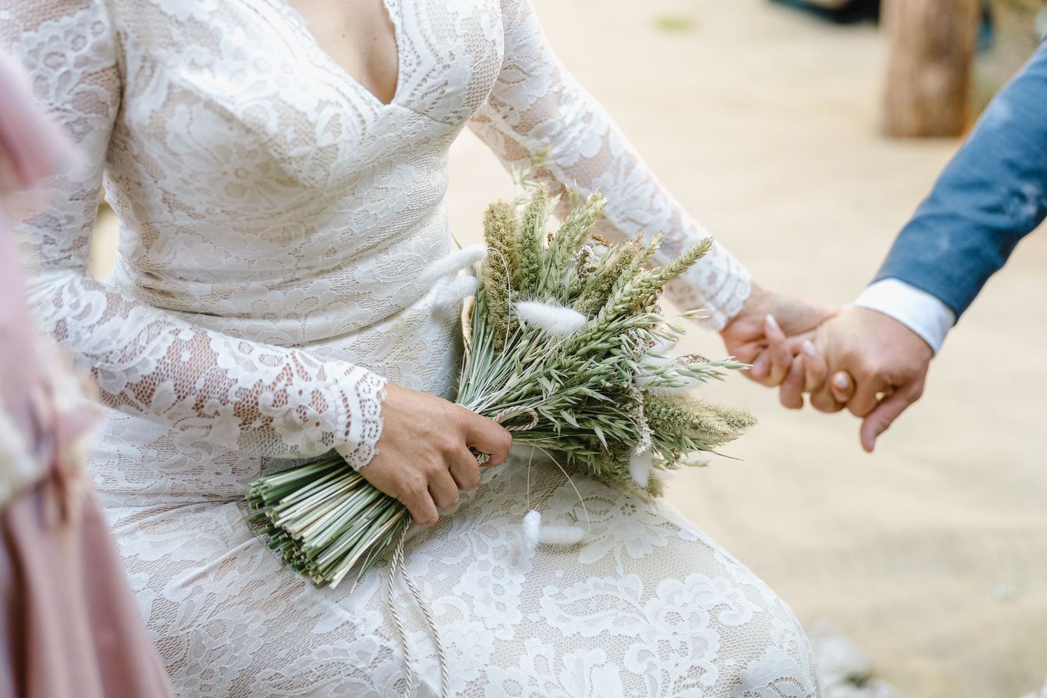 Close-up view of a bride in a lace wedding dress holding a rustic bouquet of dried wheat and greenery.