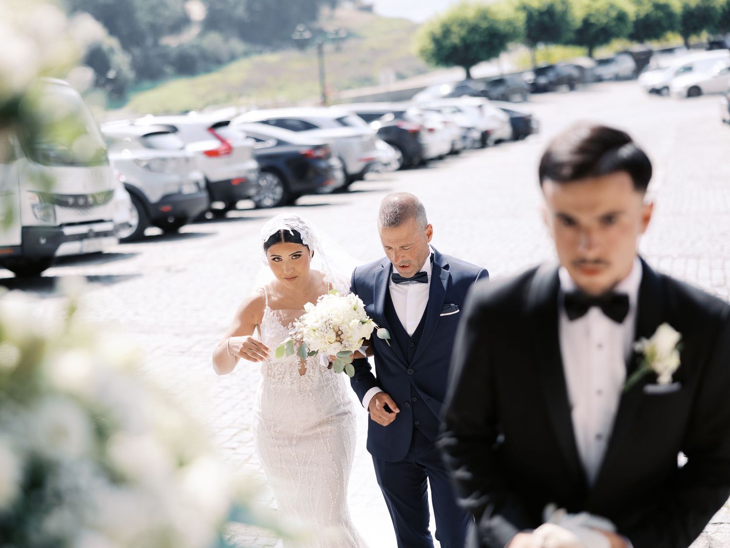 A group of formally dressed wedding guests walking and laughing together in a parking lot on a sunny day.