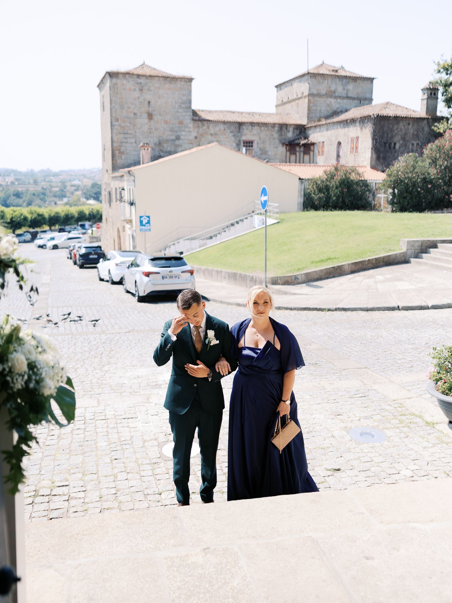 Two people in formal attire walk up a cobblestone path towards a historic stone castle building in Italy.