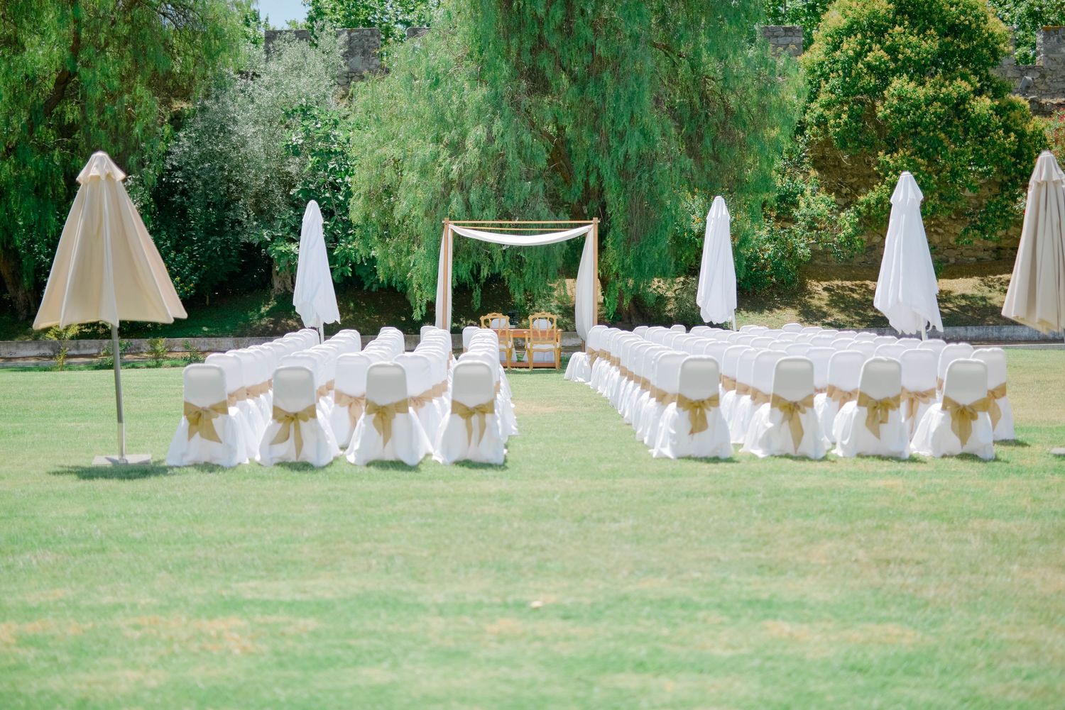 Cadeiras para a cerimônia de casamento dispostas em relvado verdejante, com decorações em tecido branco sob árvores. Evora Alentejo.