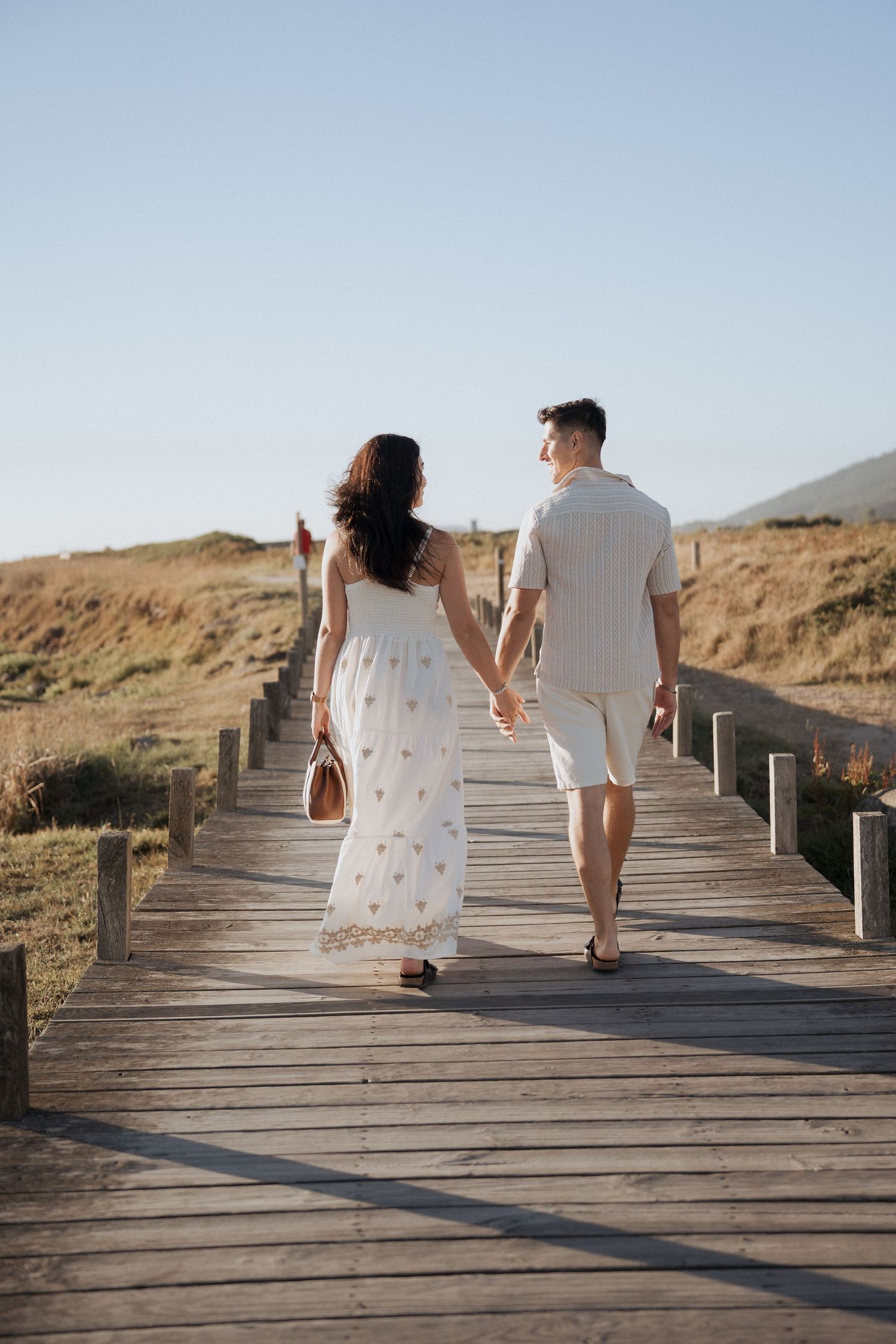 Um casal com roupas brancas combinando caminha por um calçadão de madeira durante a hora dourada. Sessão de namoro na praia