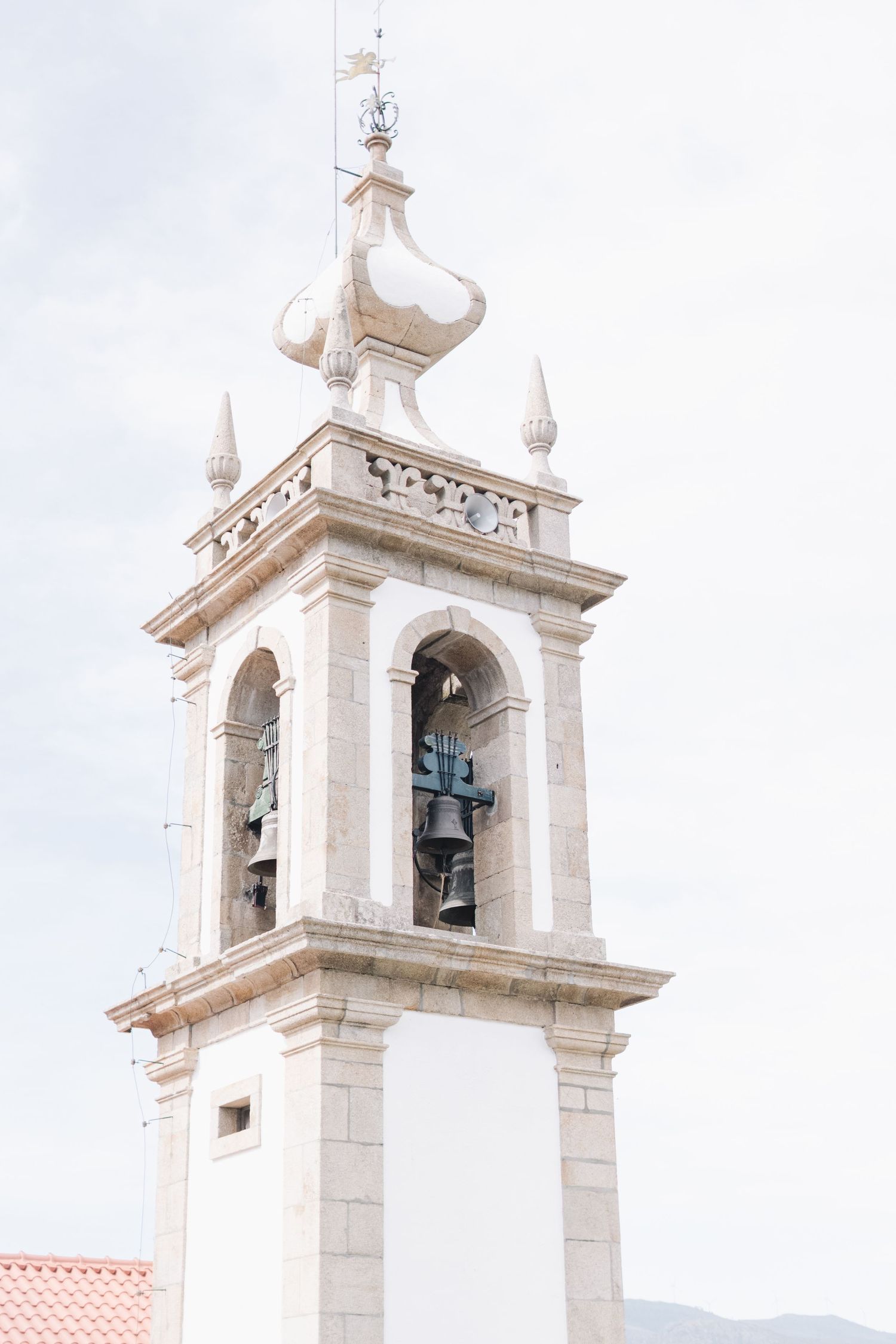 White church bell tower with ornate architectural details and bronze bells against a cloudy sky.