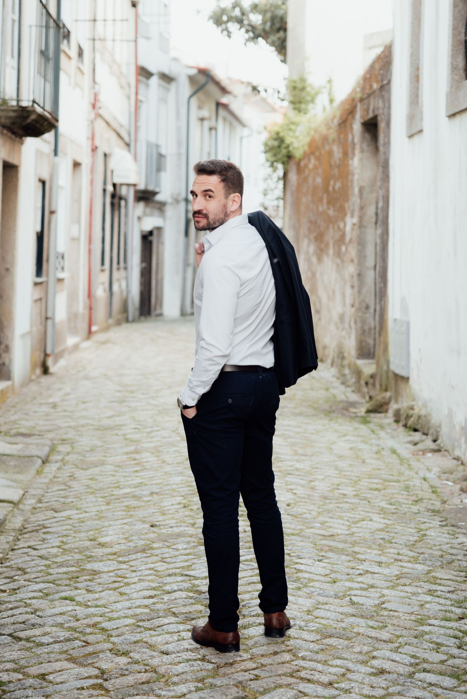 Um homem em traje formal olha para trás enquanto está parado em uma rua de paralelepípedos. Sessão fotografica de namoro na cidae