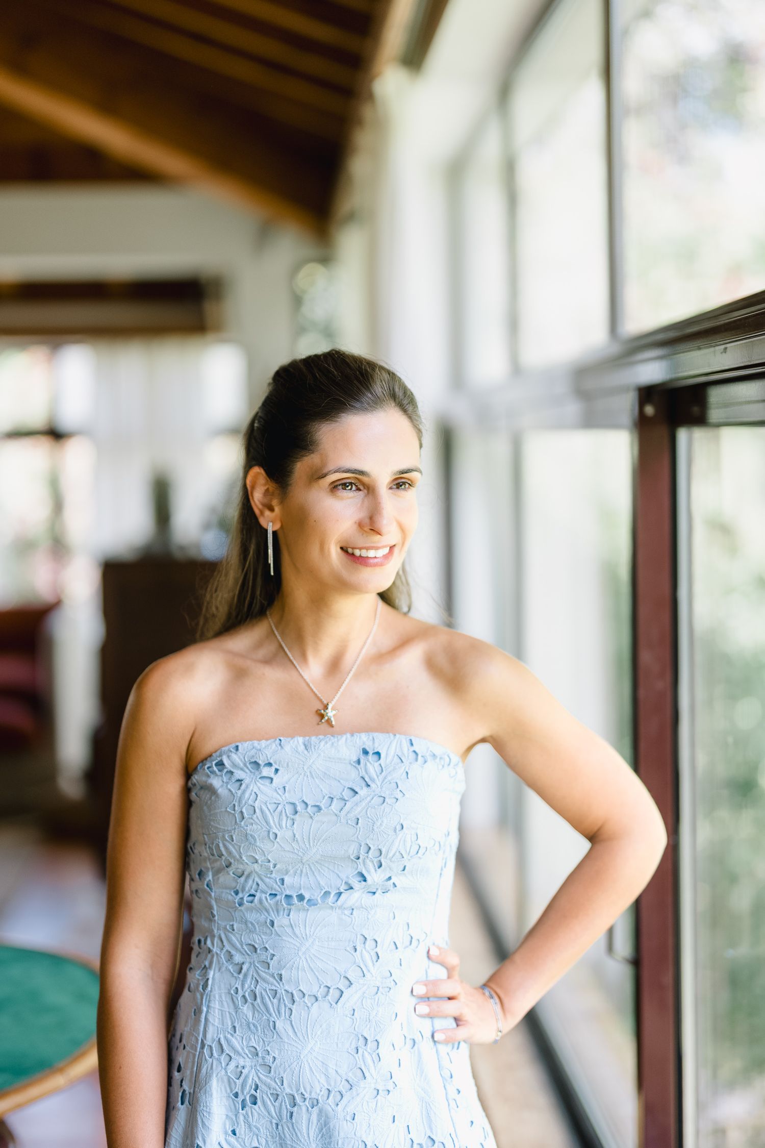 A person in a strapless light blue lace dress stands by a window in a sunlit room.