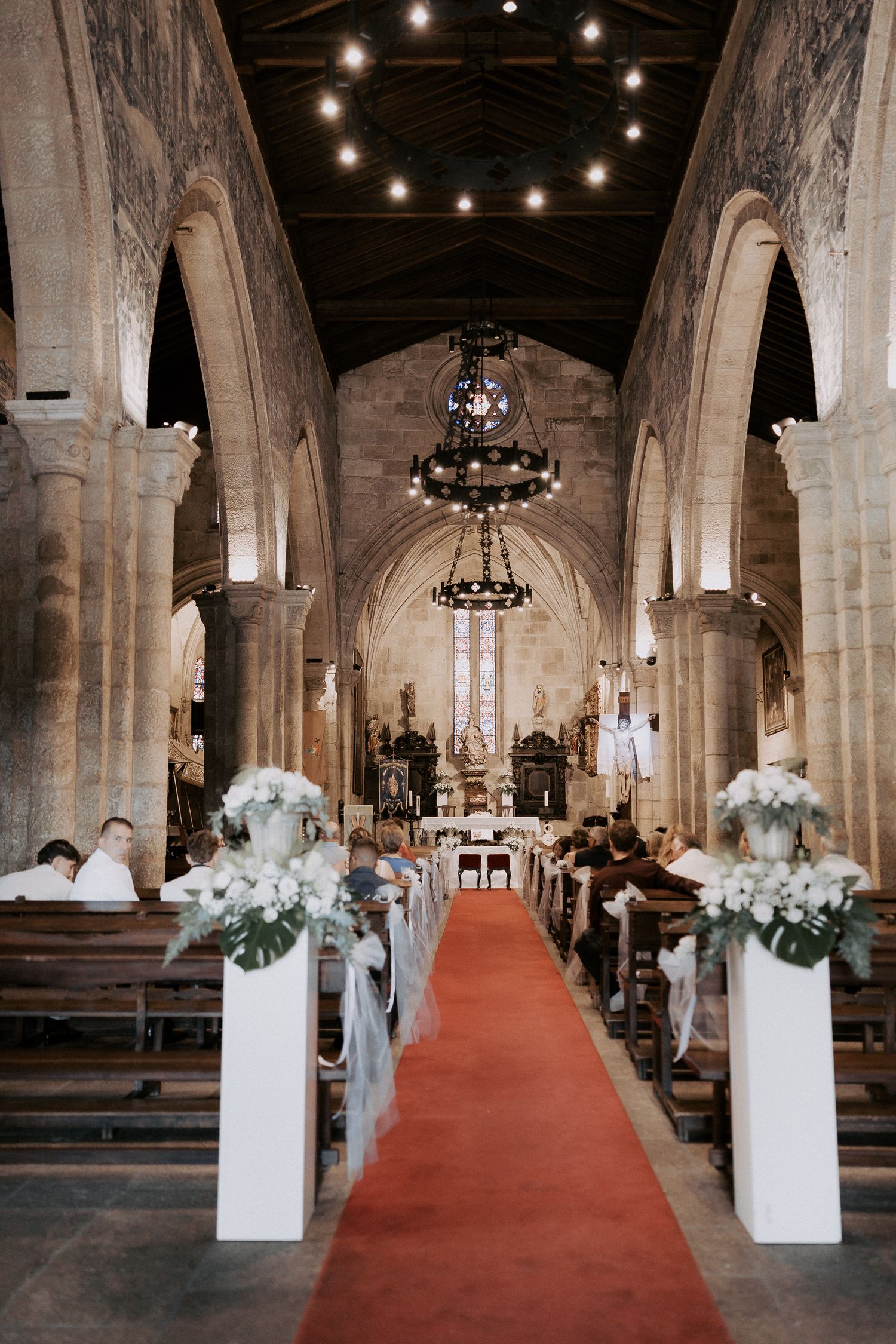 Stone church interior decorated for wedding ceremony with white floral arrangements and red carpet aisle runner.
