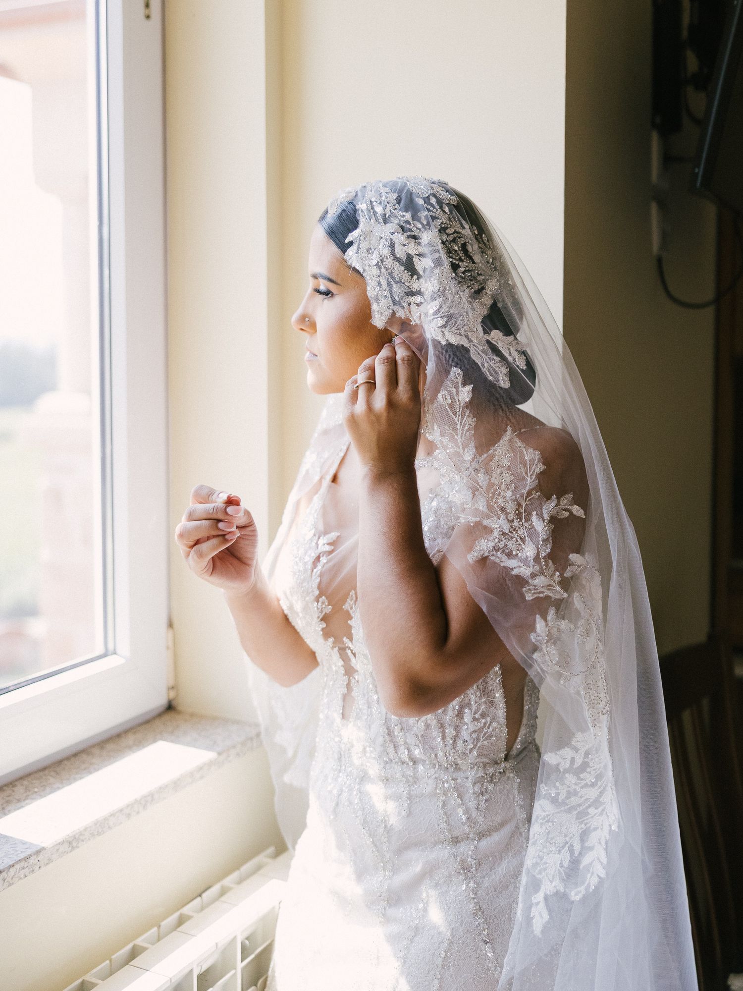 A bride in an elegant white lace wedding gown and veil stands by a window in soft natural light.