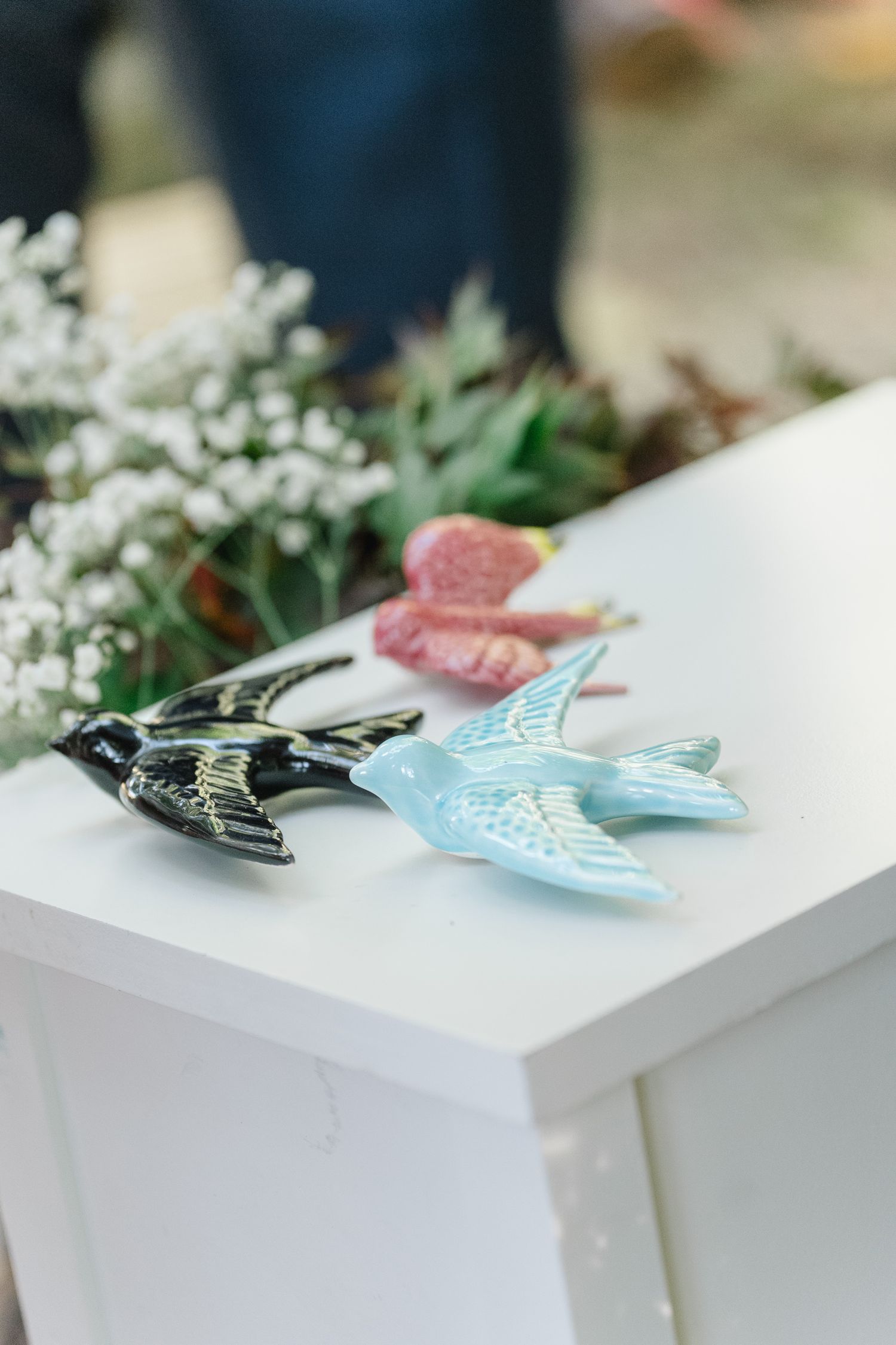 Four sequential photos showing a decorative blue bird pin placed on white surfaces with greenery in the background.