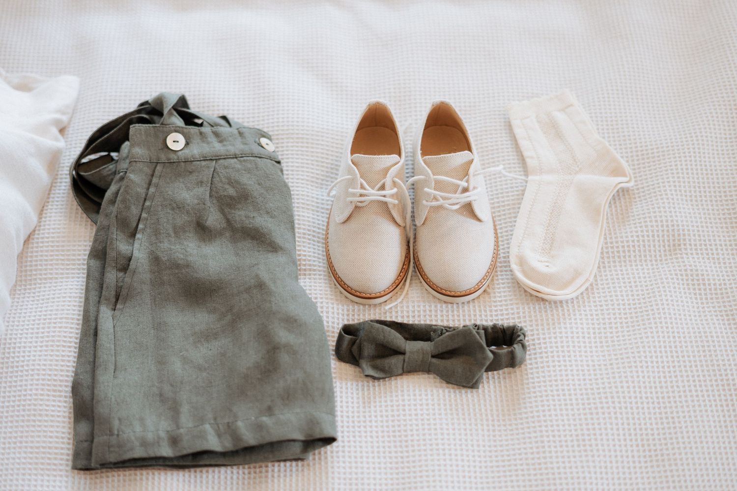 Sage green linen baby clothing and accessories including rompers, shoes, and a backpack laid flat on white fabric.