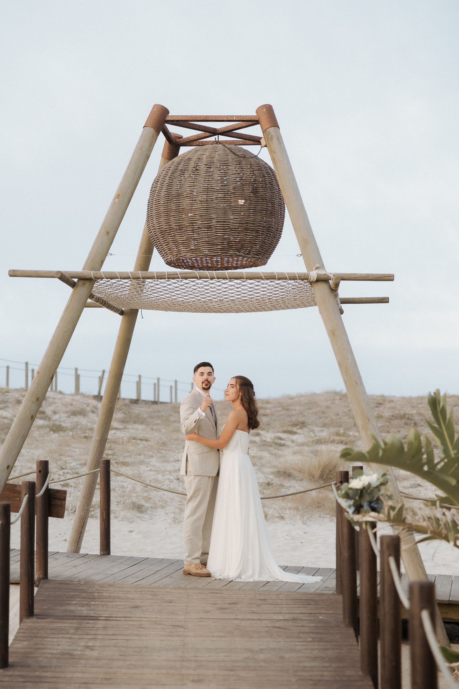 Bride and groom stand under a wicker sphere sculpture on a wooden boardwalk at a beach wedding.