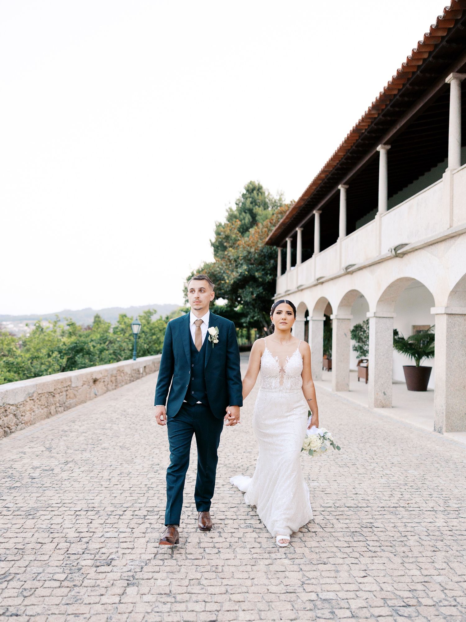 Couple walking along a scenic stone pathway with white arched colonnade in elegant wedding attire.