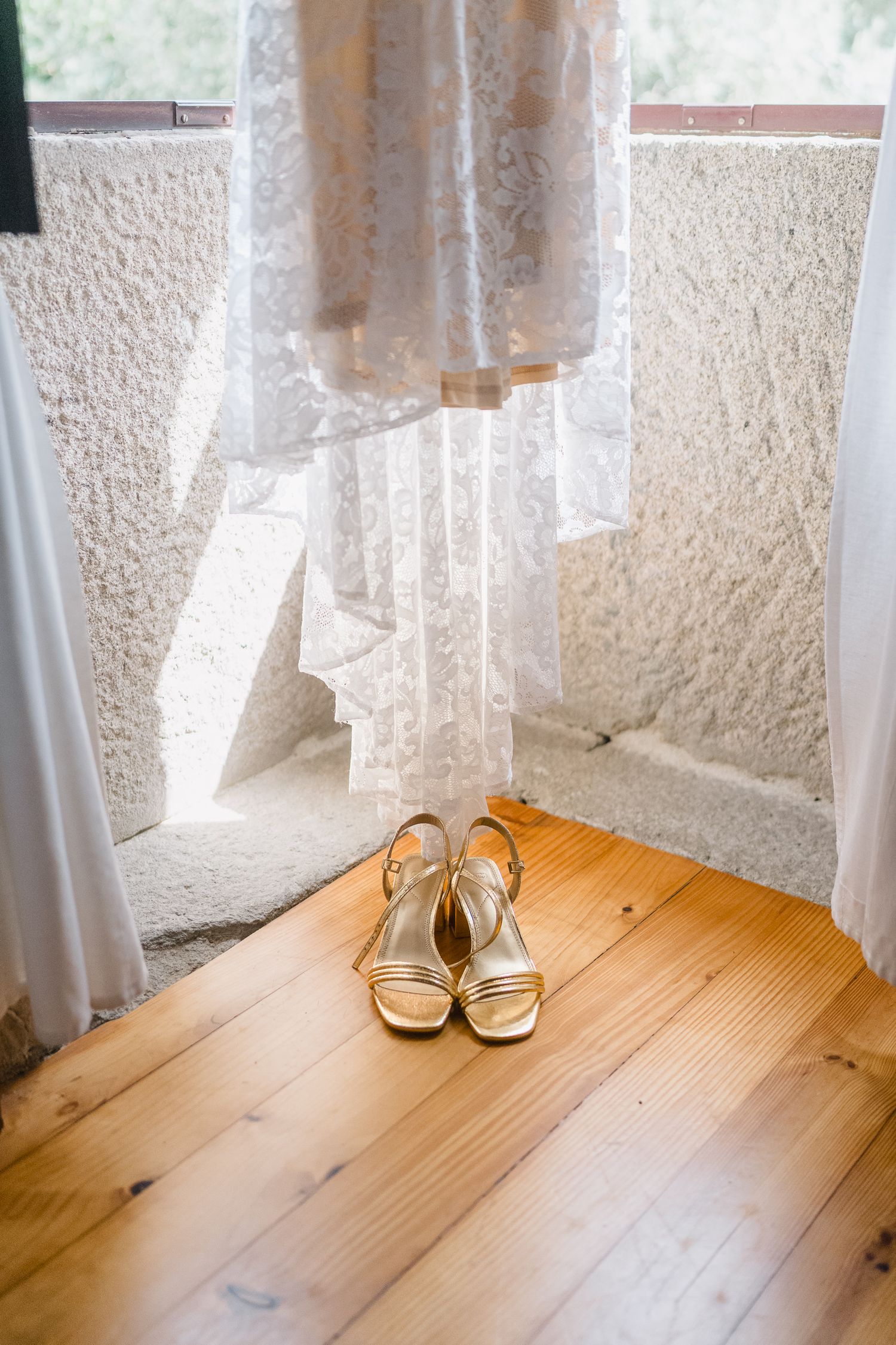 Elegant white bridal sandals on wooden floor with sheer lace curtains and sunlight streaming through window.