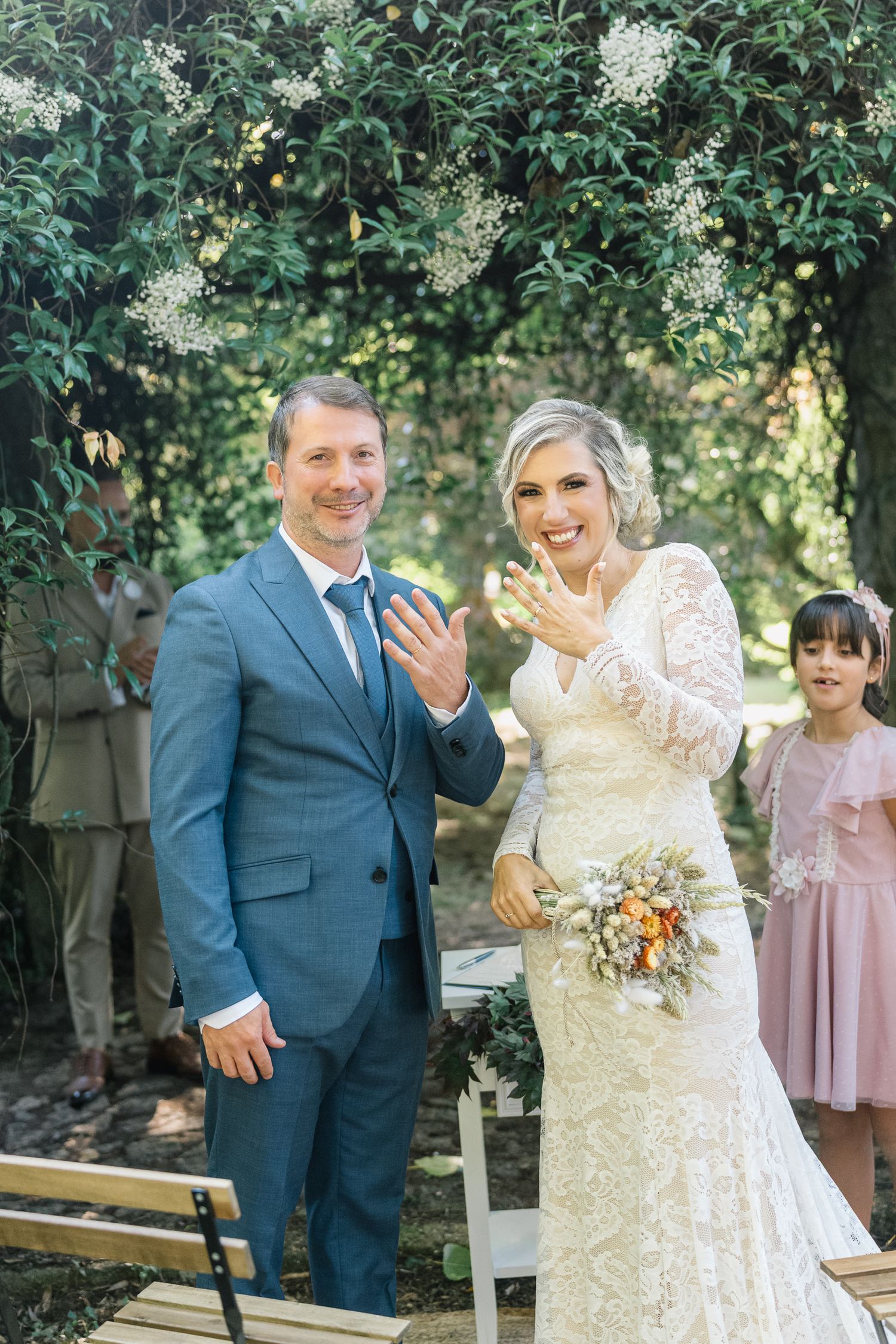 Um radiante casal de noivos partilha momentos de riso espontâneo num jardim ao ar livre, vestindo um fato azul e um vestido de renda.