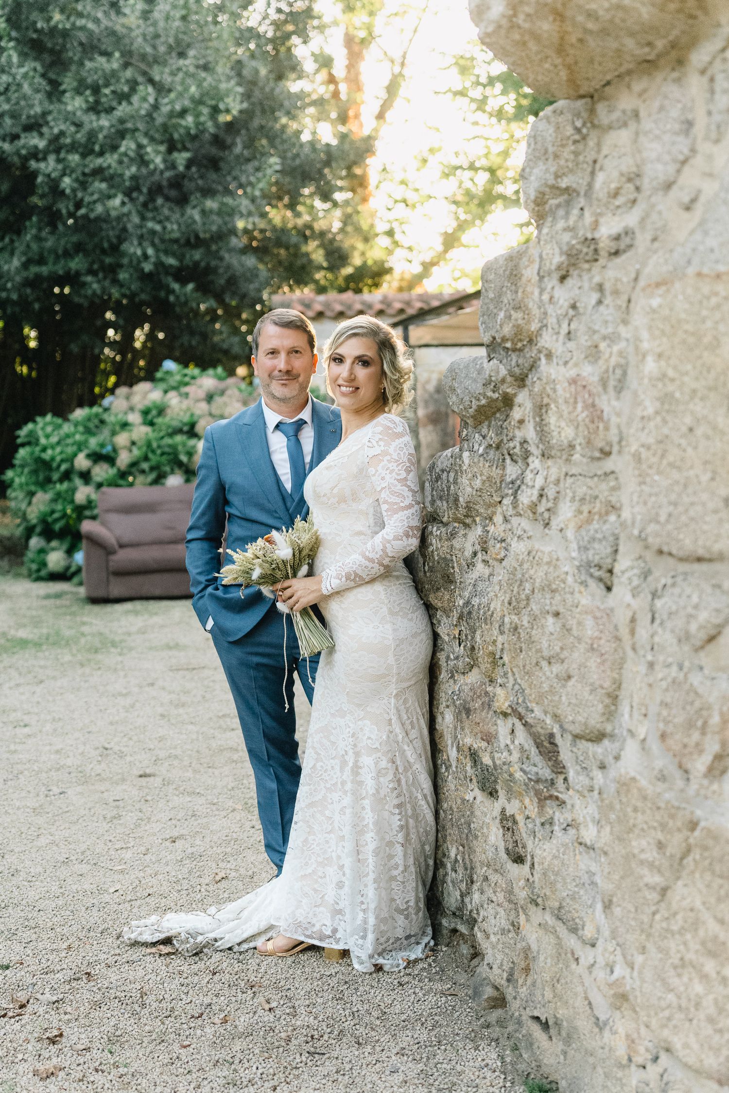 Um casal de noivos posa junto contra uma parede de pedra rústica num jardim ao ar livre. Quinta em Ponte de lima.