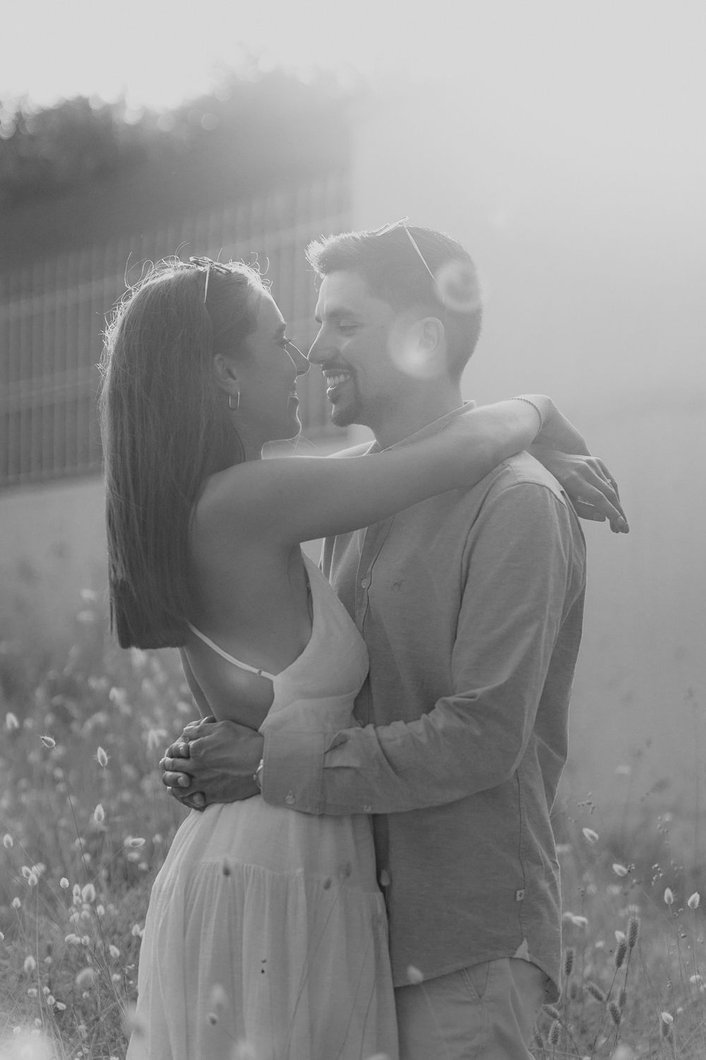 Foto romântica a preto e branco de um casal se abraçando em um prado ensolarado com flores silvestres.