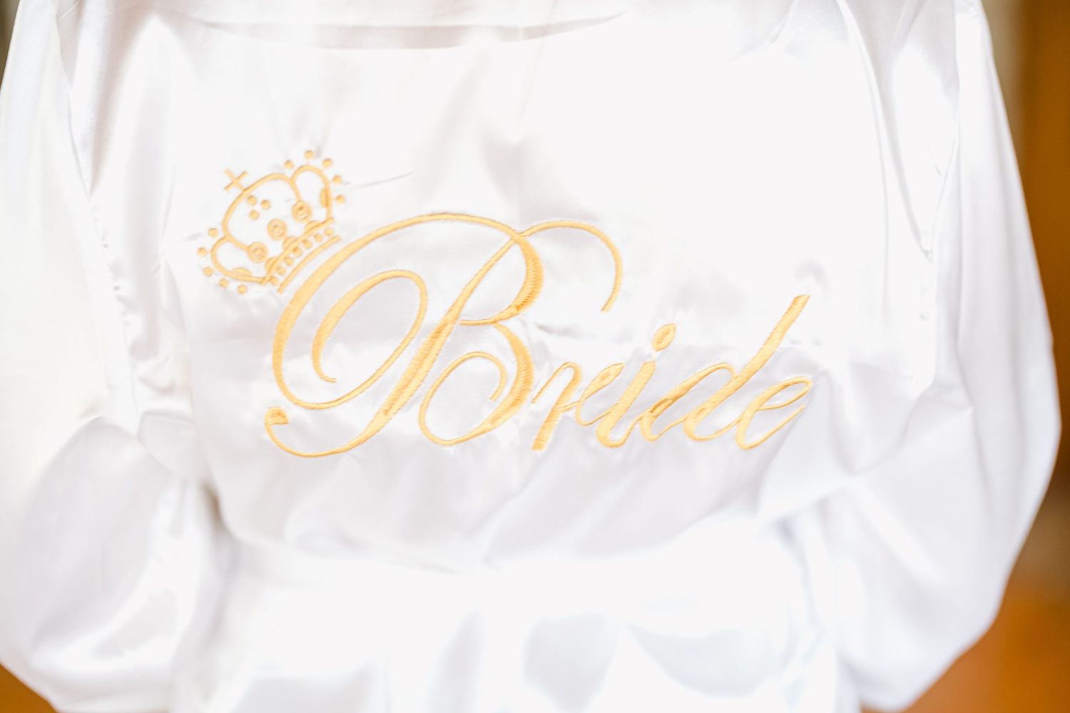 White satin robes with gold 'Bride' text embroidery hanging on wooden hangers in a row.