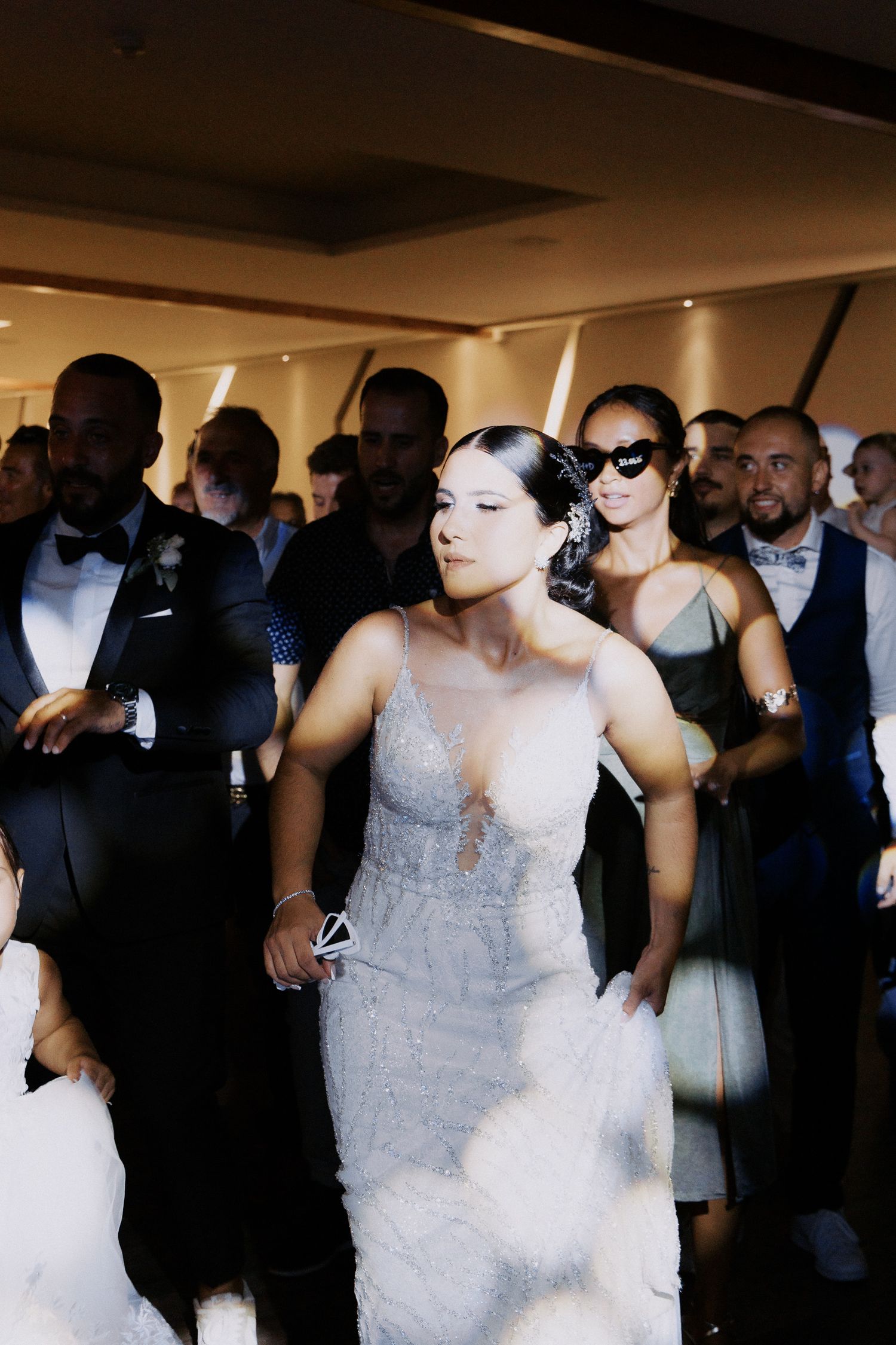 Wedding guests dance together at a reception in a dimly lit venue with the bride wearing a white lace gown.