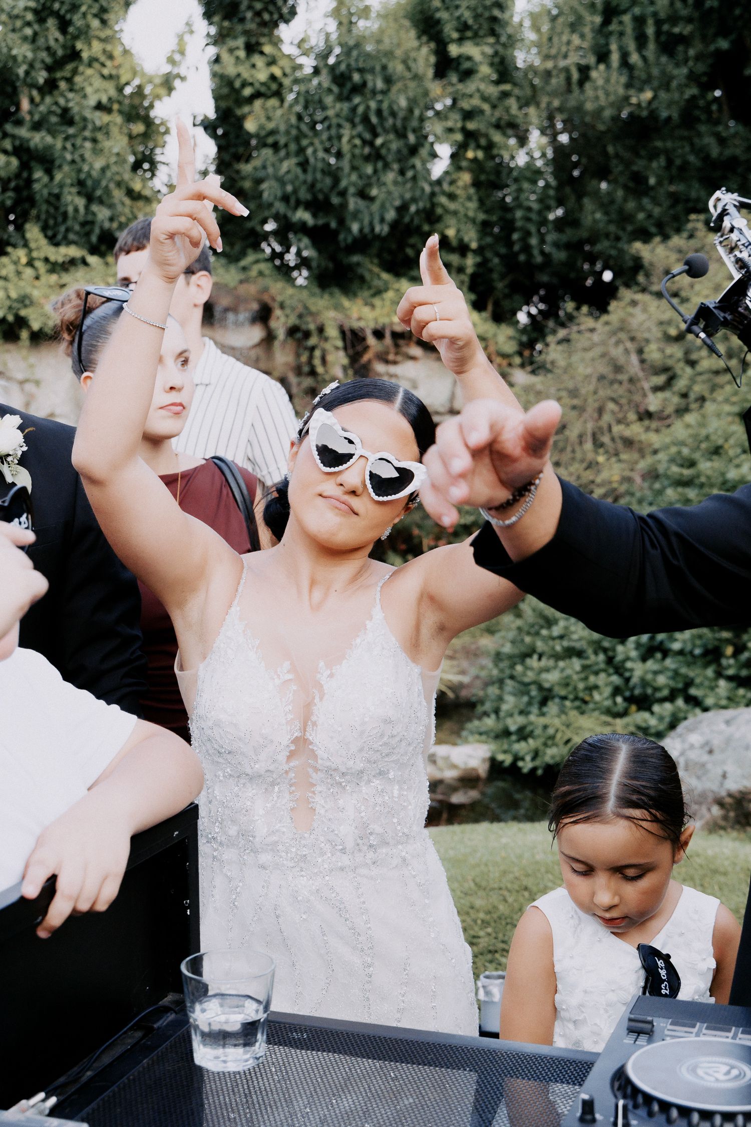 A group of wedding guests in black and white attire raise their hands in celebration at an outdoor reception.