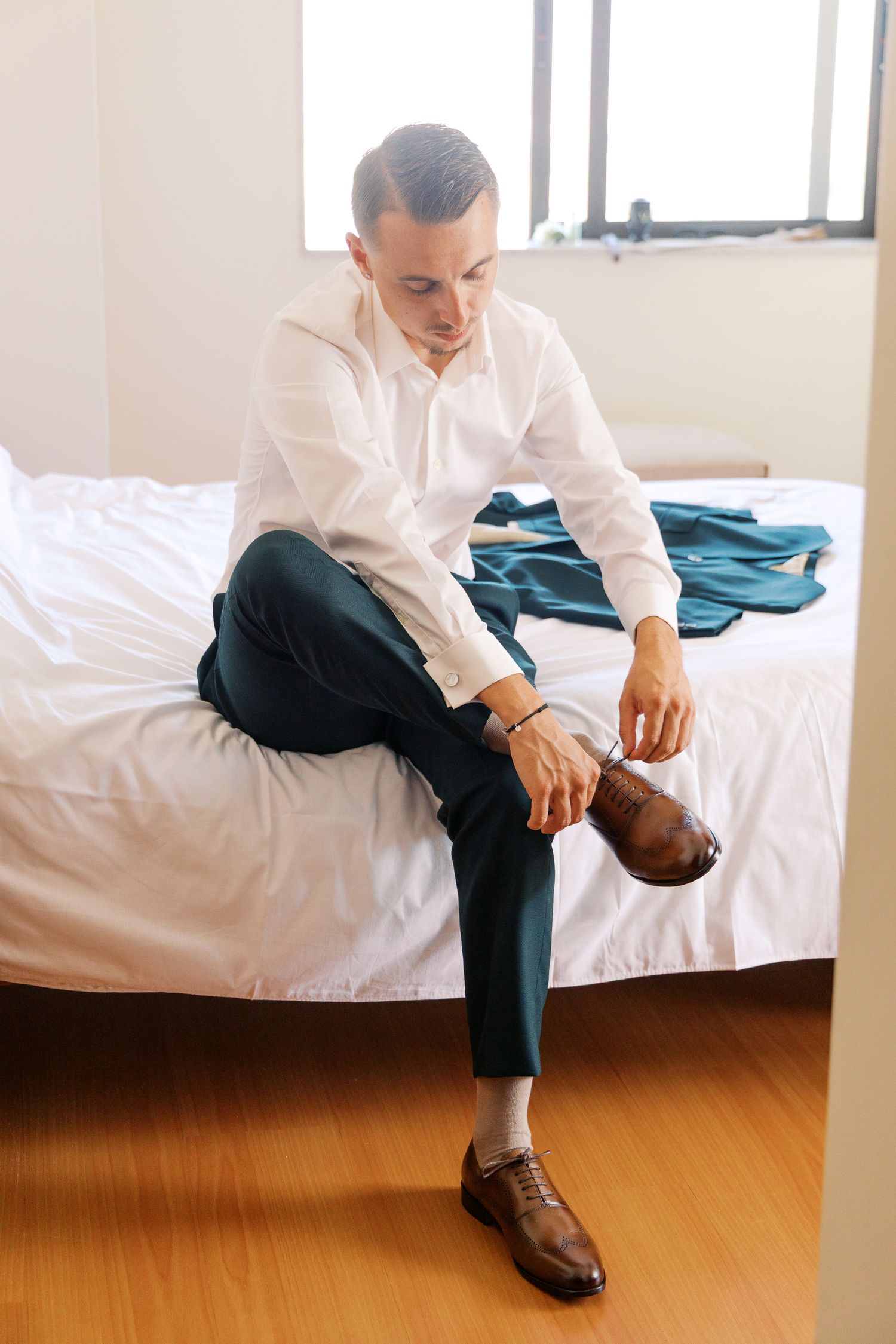Person in white shirt and dark pants sits contemplatively on white bedding in a minimalist room setting.