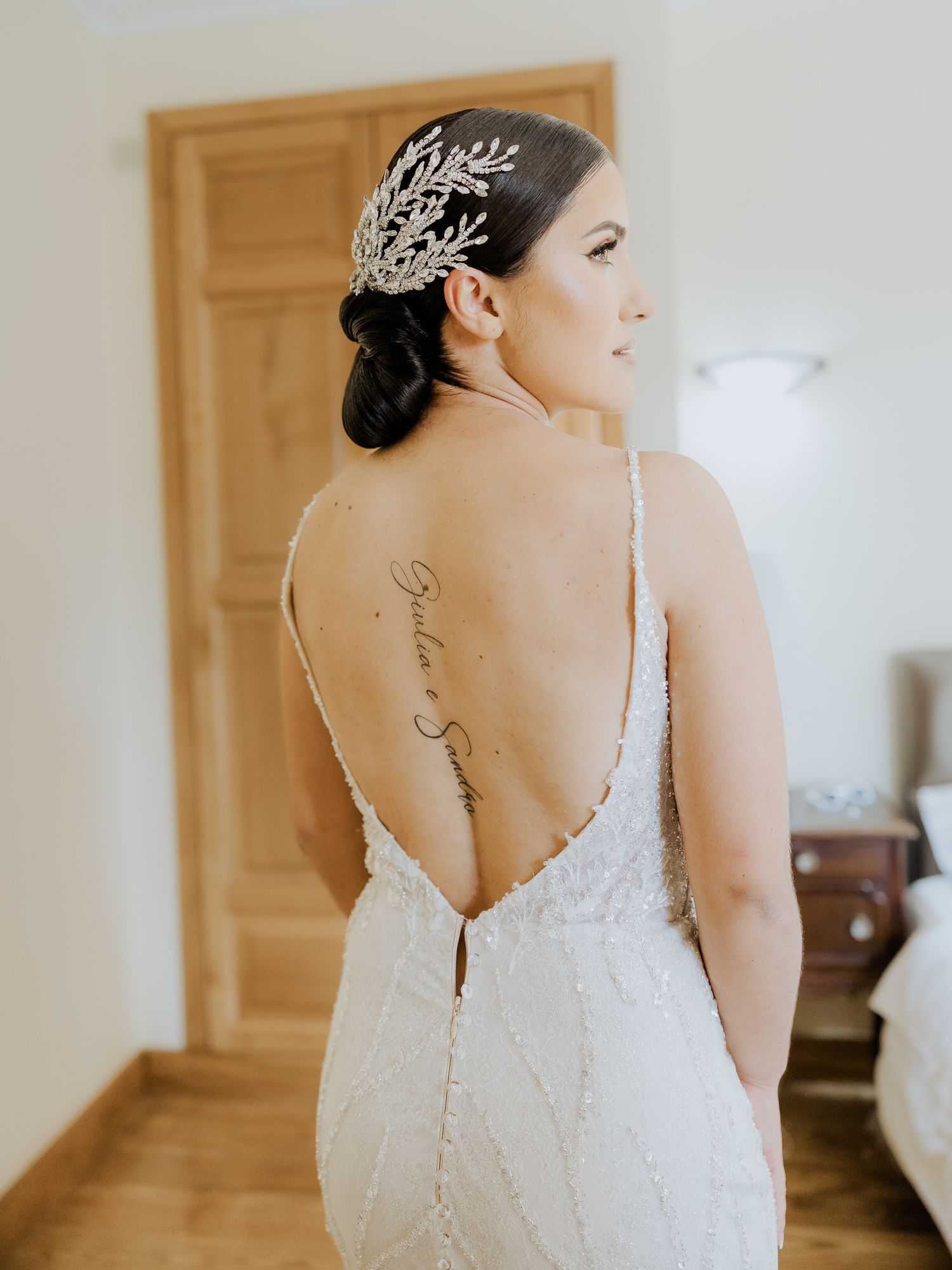 A bride in a white lace gown with a crystal headpiece shows off her back tattoo and elegant low-back dress detail.