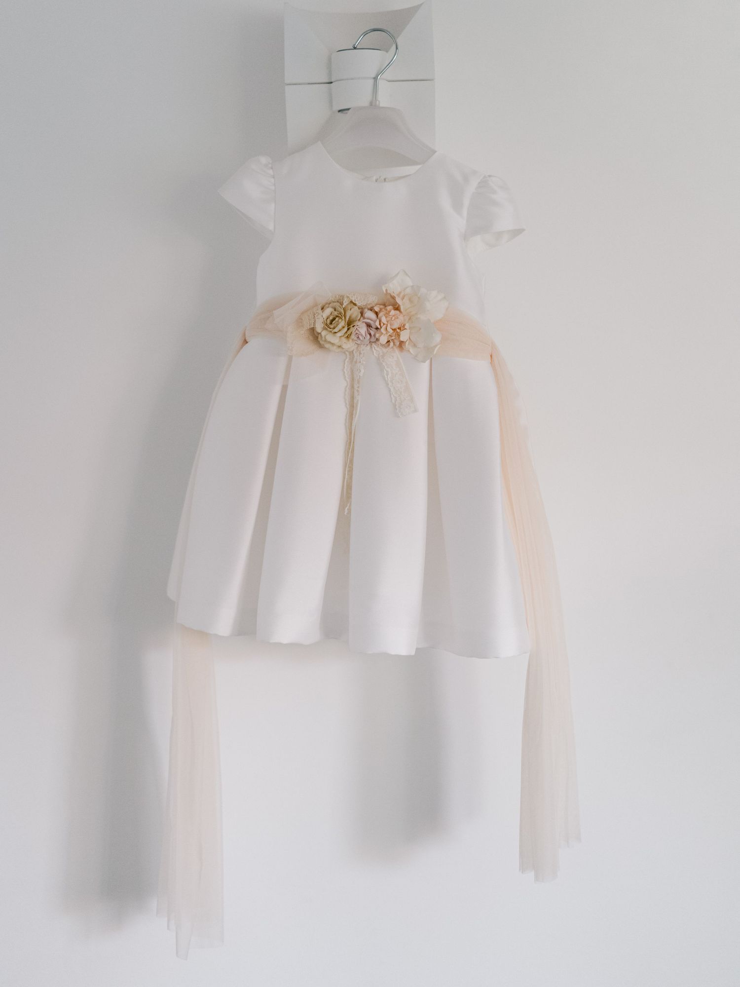 A series of delicate white baby dresses with ruffled details and soft peach accents hanging against a white wall.
