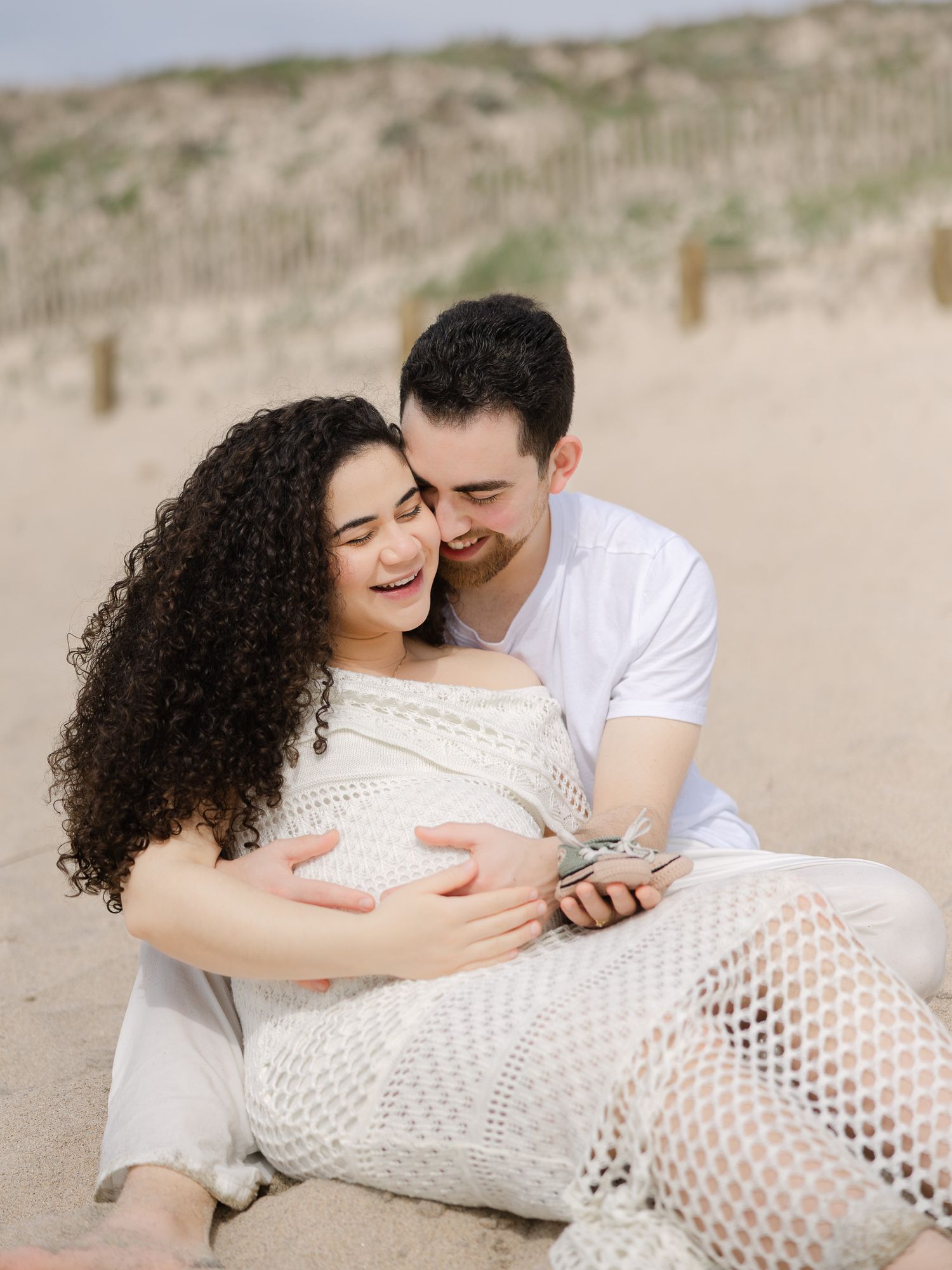 A couple sits on sandy beach, woman in white lace dress cradling baby bump, man embracing her from behind, both smiling.