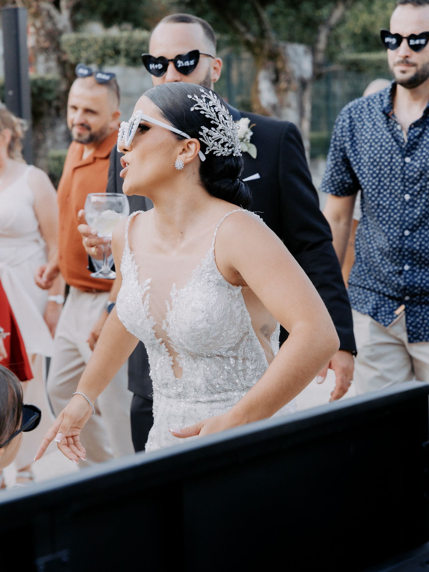 A bride in a white lace wedding dress and crystal headpiece dances with guests at an outdoor reception.