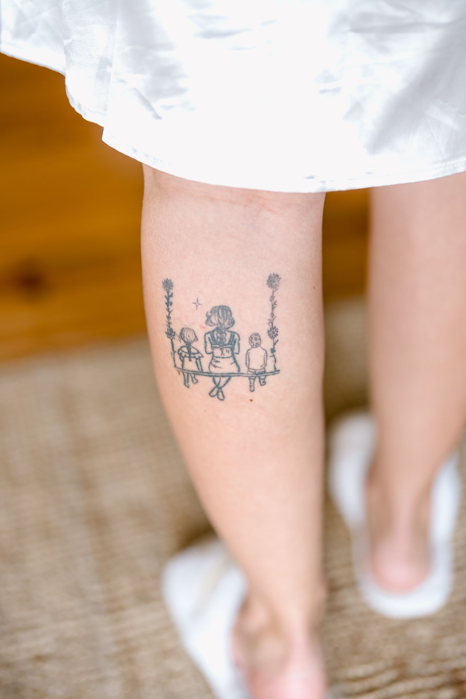 A delicate tattoo on a leg showing a whimsical design of figures sitting on a swing set in black ink.