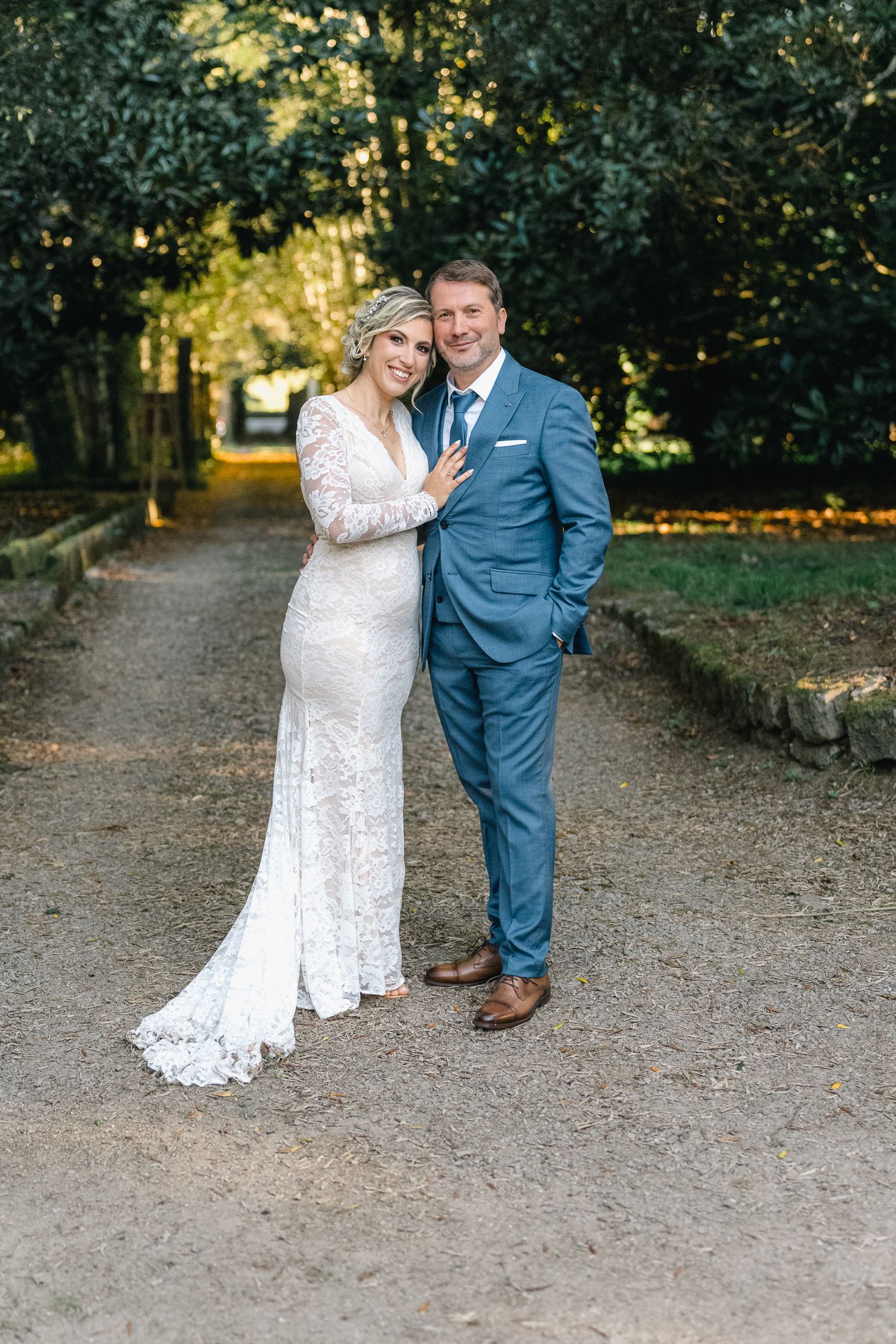 Um casal posa junto num caminho de cascalho, rodeados por uma exuberante folhagem de outono, vestindo as suas roupas de casamento.