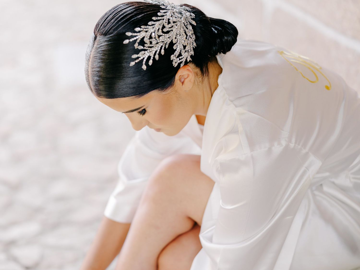 A bride in a white dress sits gracefully on a bed with soft lighting and delicate details.
