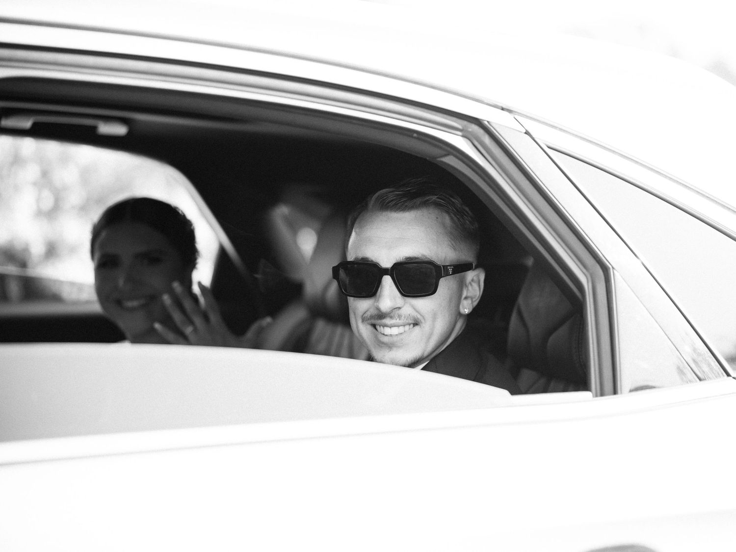 A car driver wearing sunglasses and a cap is seen through the driver's side window of a vehicle.
