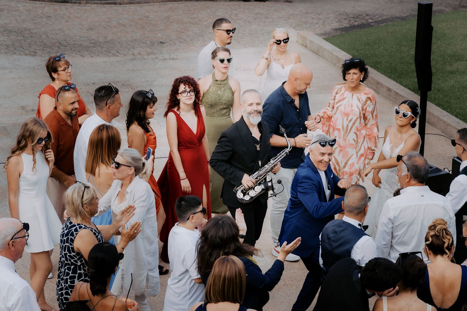 A group of wedding guests gather outdoors during a summer celebration with people in colorful dresses and formal attire.