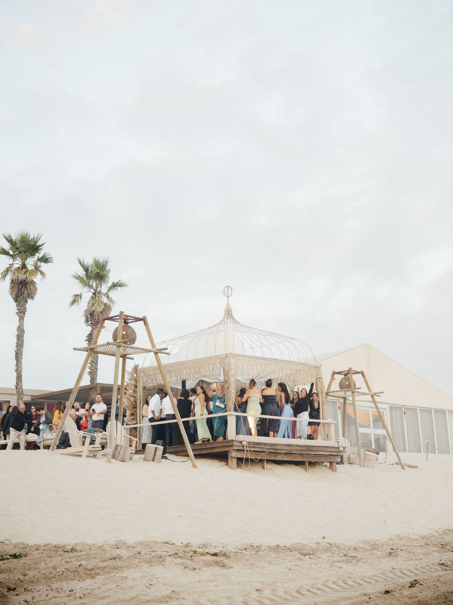 White beach wedding tent setup with palm trees and guests gathering on a sandy shoreline during a sunny day.