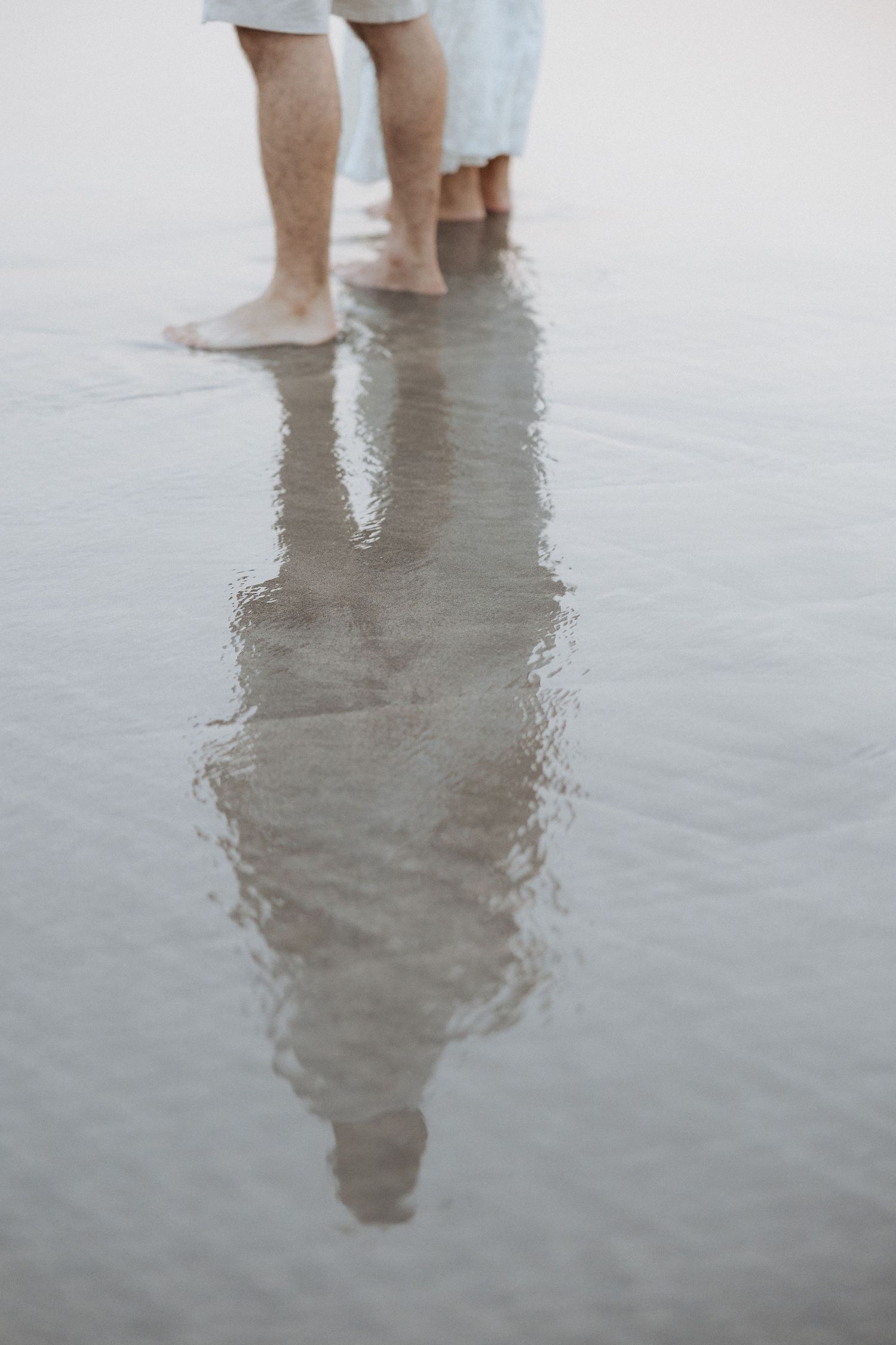 Pés descalços refletidos na água rasa criam um efeito espelhado. Sessão fotográfica de namoro na praia