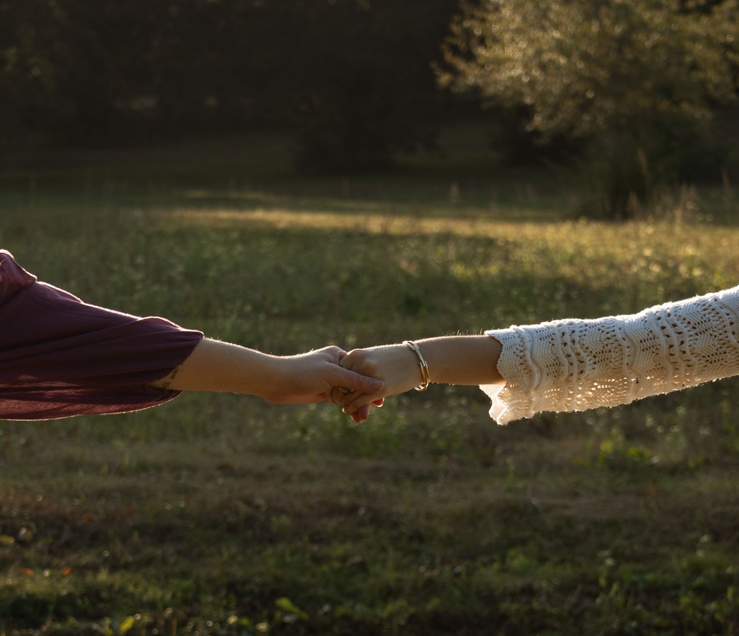 Two people holding hands at arms length in a sunlit green meadow during golden hour.