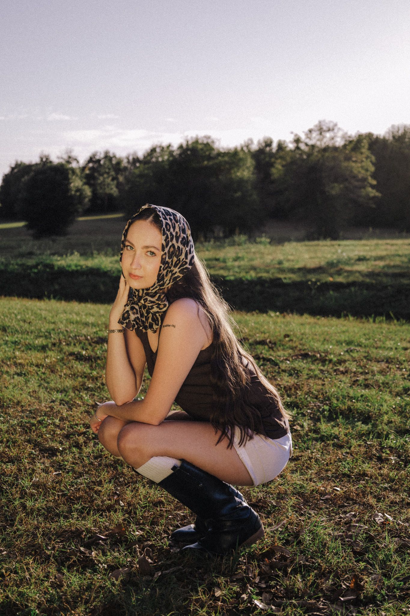 Young woman in leopard print headscarf and black boots posing in green field at golden hour.