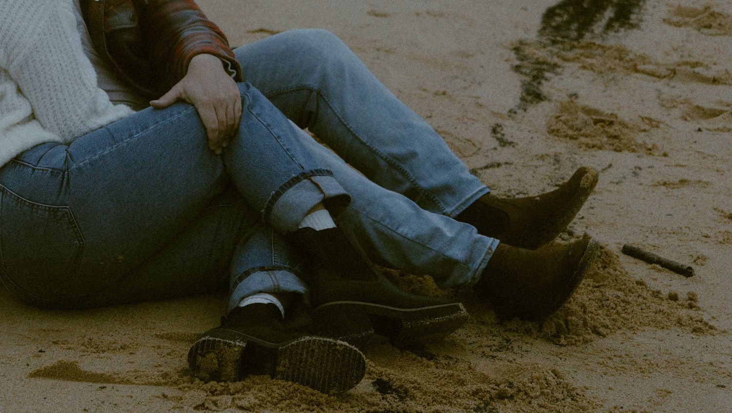 Couple sitting together on sandy beach, wearing jeans, with boots and sneakers visible in natural light.